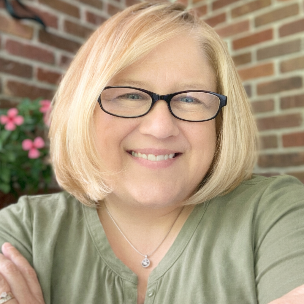 Close-up of a smiling woman with blonde hair, wearing glasses, a green top, and a necklace, standing in front of a brick wall with pink flowers in the background.