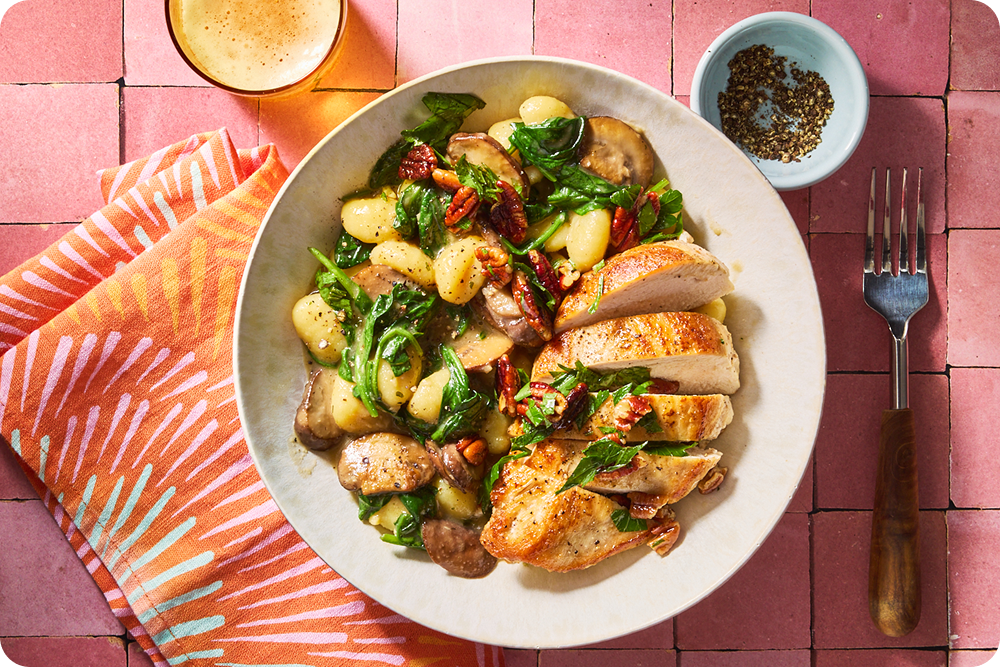 Sliced seared chicken breast next to gnocchi with sautéed spinach and mushrooms, topped with chopped pecans in a white bowl over a pink tiled background.