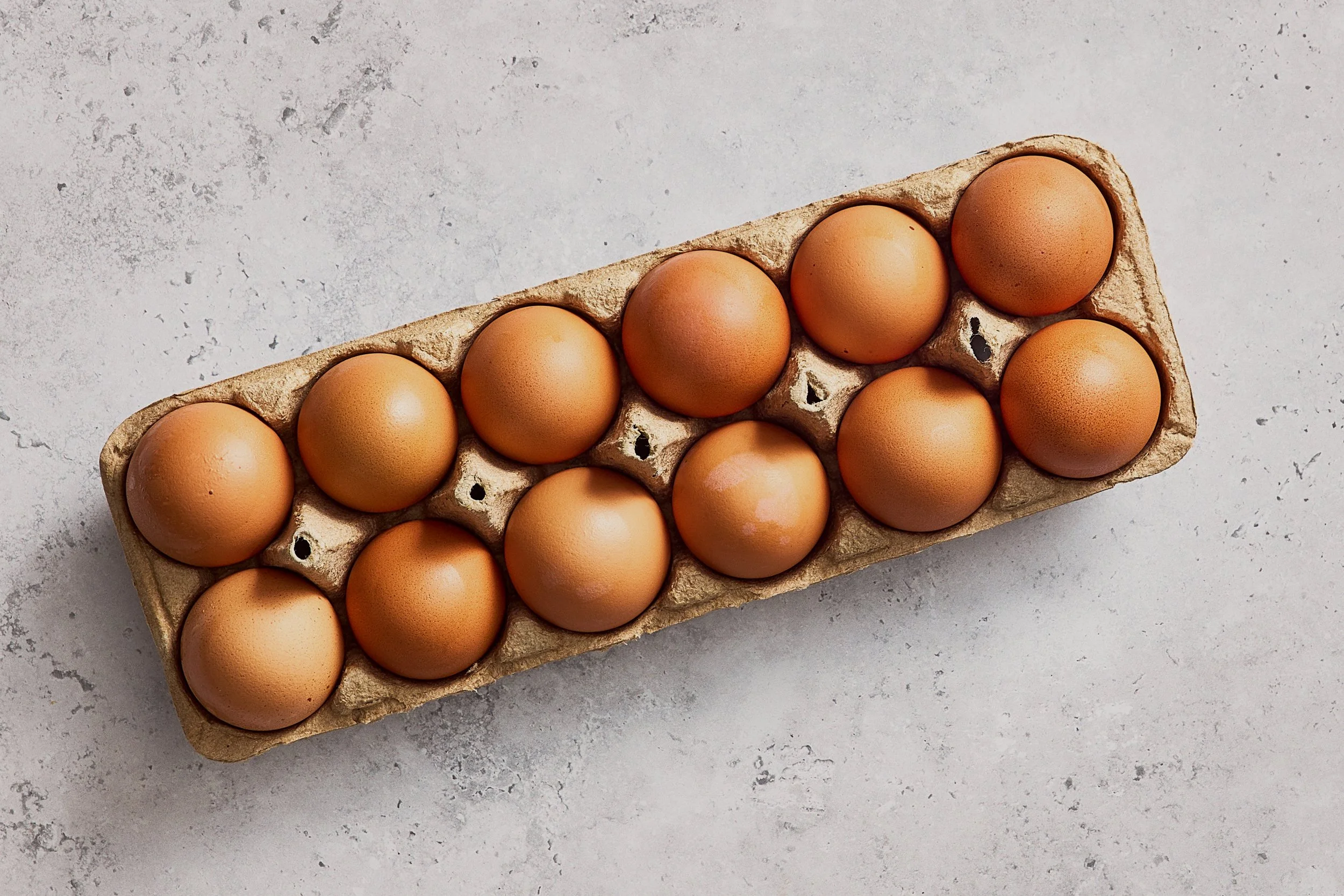 Flat-lay photograph of a full cardboard carton with a dozen brown eggs arranged in two rows on a textured stone countertop.