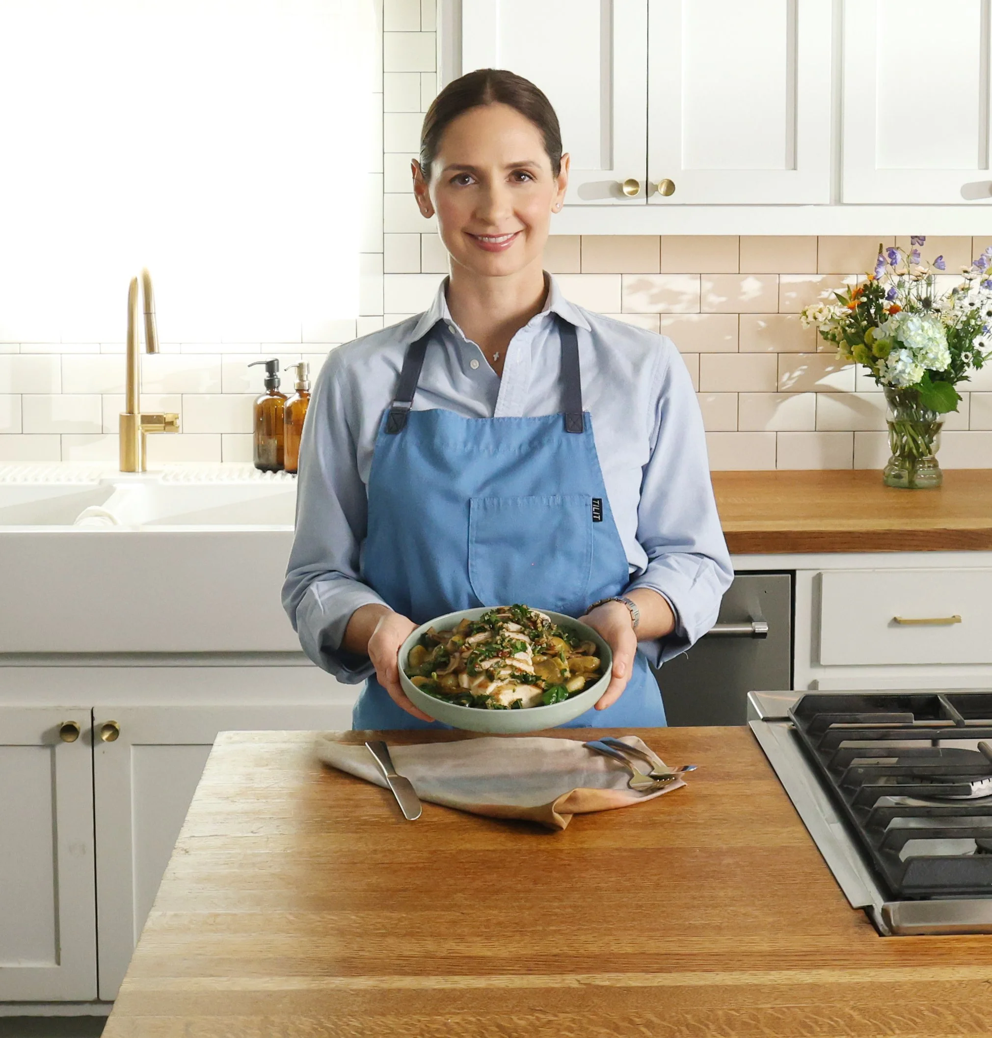 Chef Tracy Malechek-Ezekiel, a smiling woman with dark hair neatly pulled back, wears a light blue shirt and matching apron while holding a bowl of chicken and gnocchi in a bright, modern kitchen.