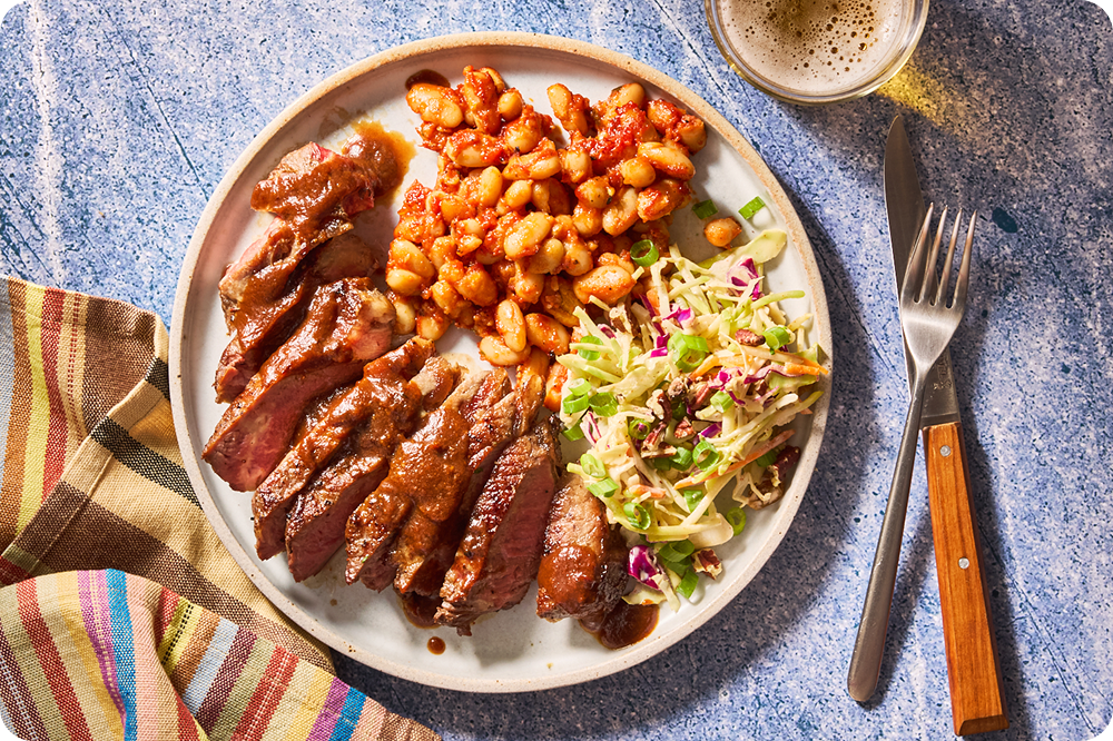 Sliced steak drizzled with savory sauce, served with tomato-stewed white beans and a crisp cabbage coleslaw on a white plate over a blue textured background.