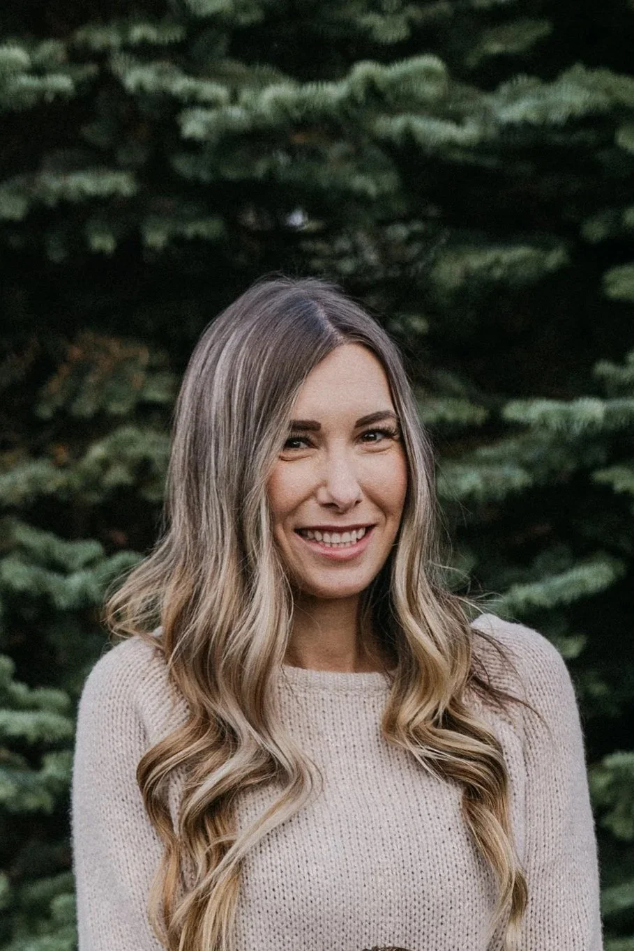 A woman with long wavy blonde hair smiling outdoors in front of green pine trees.
