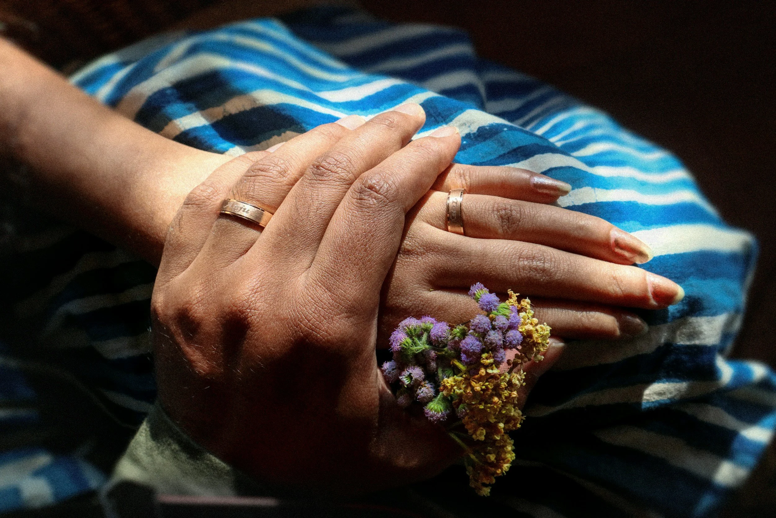 Two hands with wedding rings resting on a striped blue and white fabric, one hand holds a small bouquet of purple and yellow flowers.