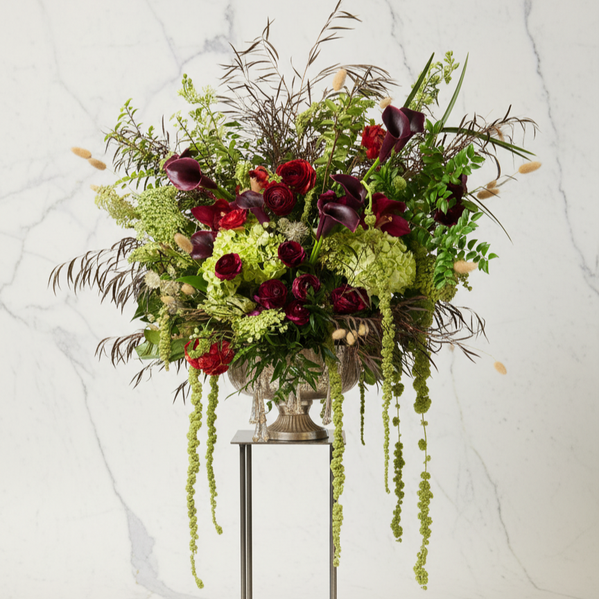 Large floral arrangement with dark purple calla lilies, red roses, green hydrangeas, and tall brown and green foliage in a silver vase on a stand against a white marble background.