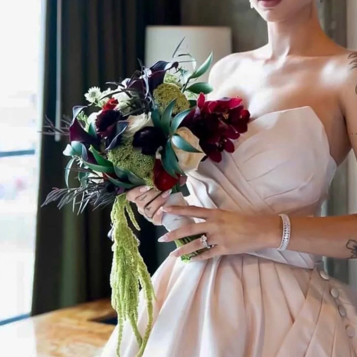 A woman in a strapless, pale pink dress holding a bouquet of dark purple, white, and green flowers, with a green ribbon hanging from it, standing indoors with a window and dark curtains in the background.