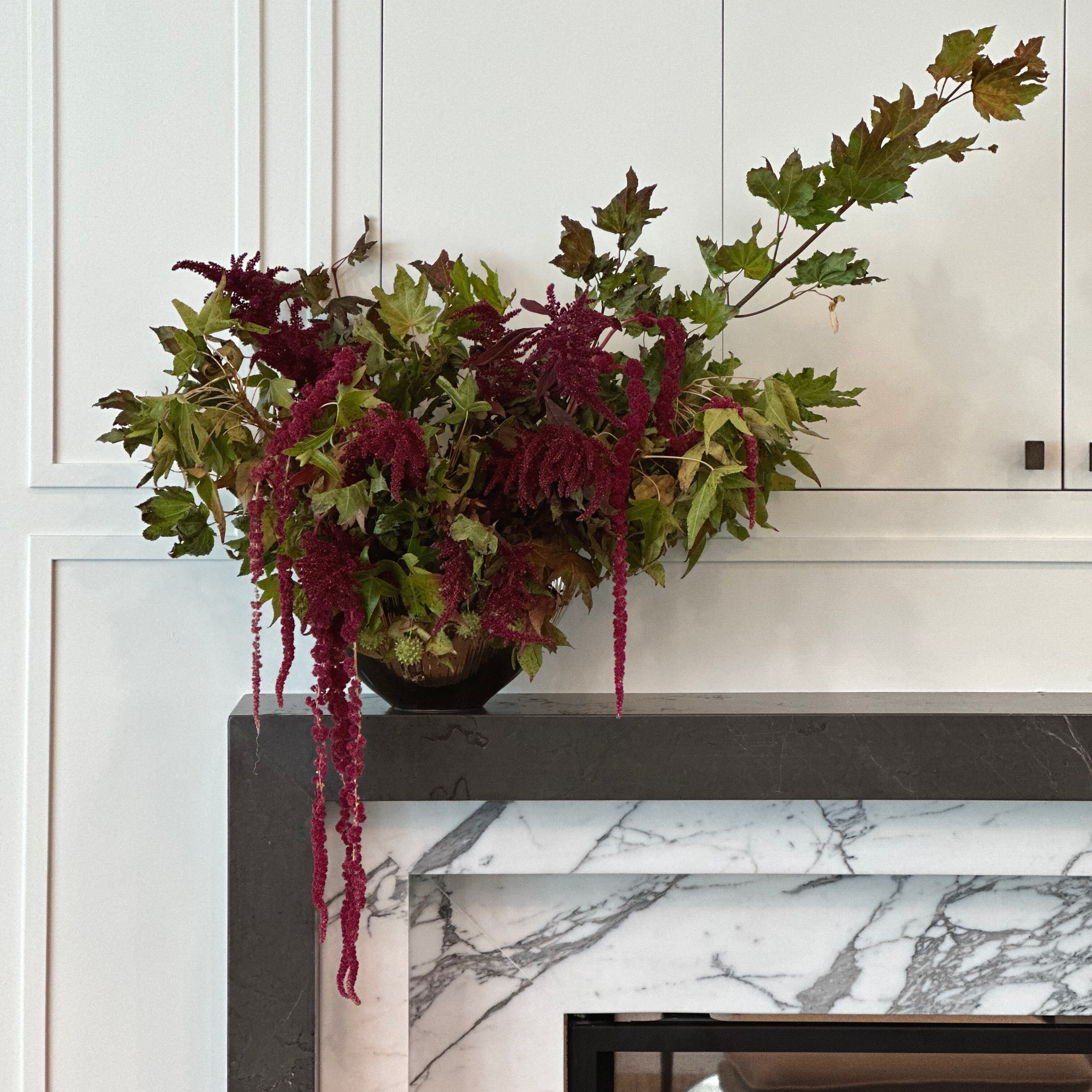 A large bouquet of maroon and green plants and flowers in a dark vase, placed on a marble and black stone fireplace mantel against a white paneled wall.