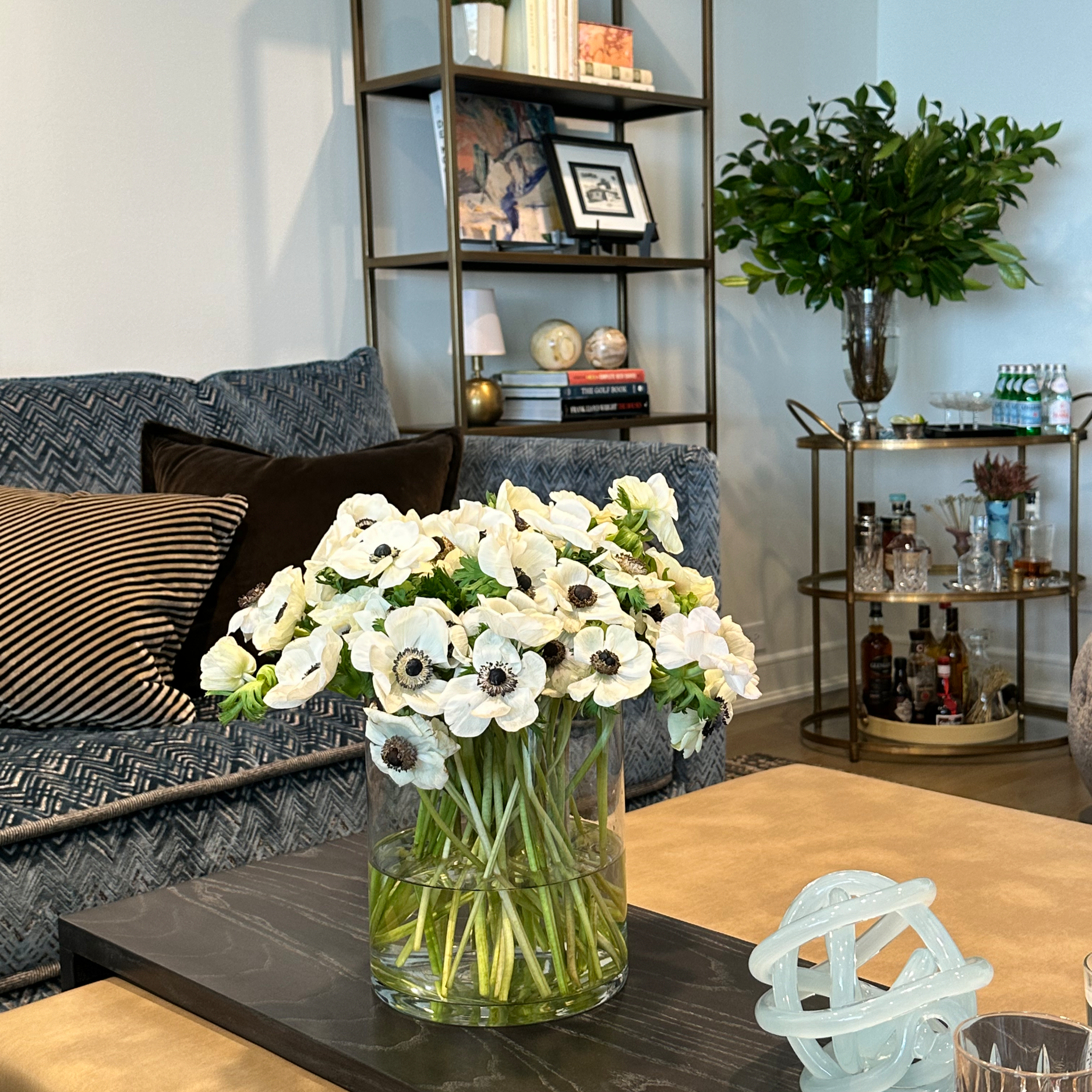 A clear glass vase filled with white flowers on a black tray, placed on a brown table in a living room.