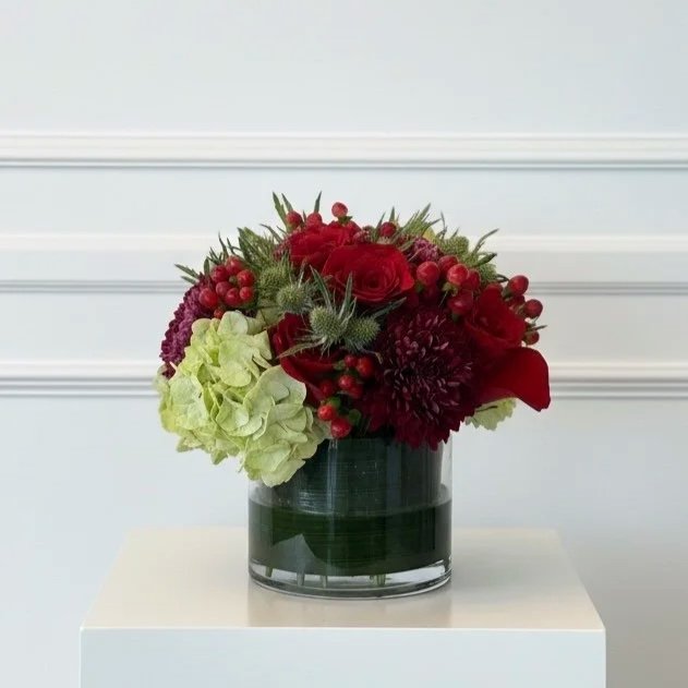 A floral arrangement in a black vase with red roses, red berries, purple flowers, green hydrangeas, and other greenery, placed on a white surface against a light background.