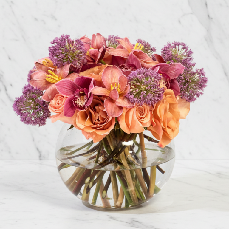 A round glass vase filled with water holding a colorful bouquet of peach roses, pink orchids, and purple allium flowers on a white marble surface.