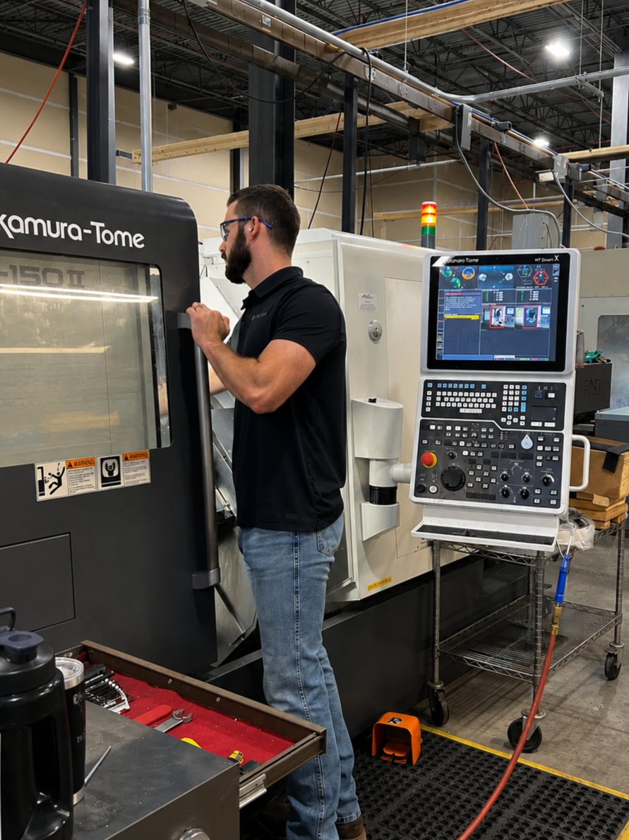 A man working on a CNC machine inside a machine shop / manufacturing facility. The man is wearing safety glasses and black shirt, with machine control panels and tools nearby.