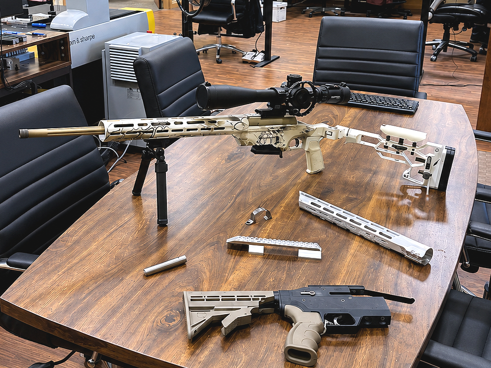 Above a wooden conference table, a sniper rifle with a scope and bipod, disassembled pistol with slide and frame, and various gun parts are arranged showcasing firearm machining experience.