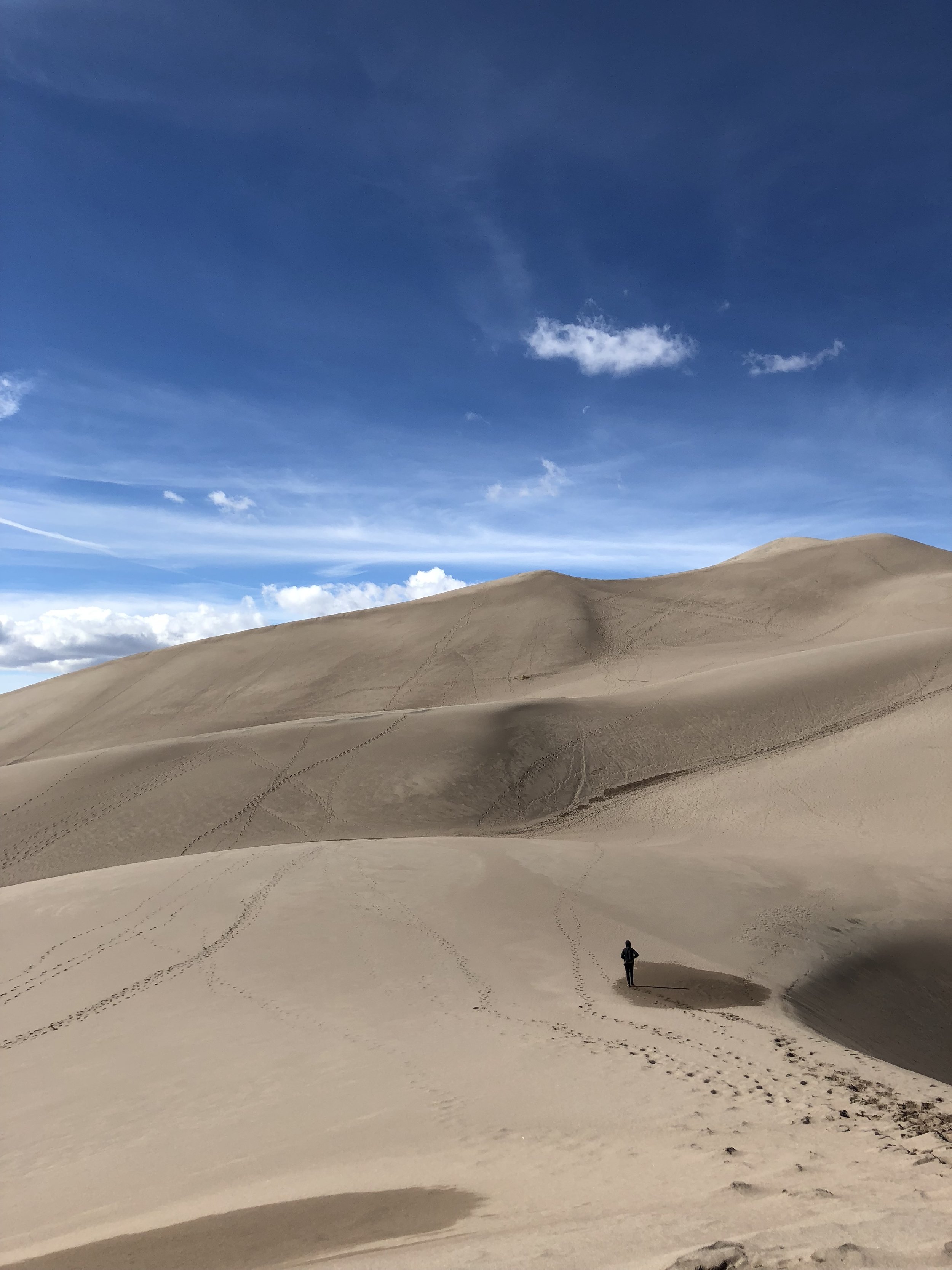 A person standing alone in a vast desert of sand dunes. The sky above is mostly clear with some clouds and a faint moon visible.