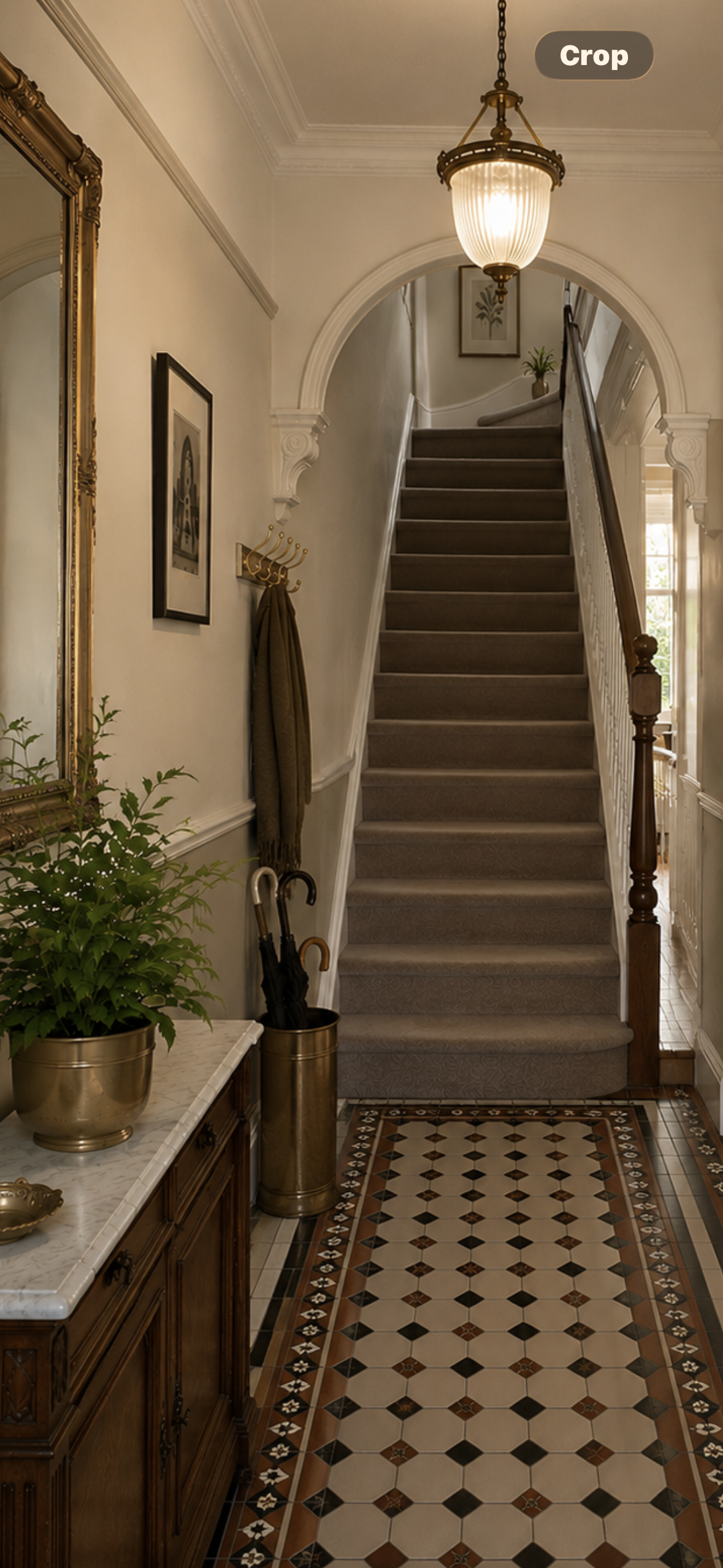 Interior view of a vintage foyer with patterned tile floor, a wooden console table with a potted plant and umbrella stand, a wall mirror, framed artwork, a coat hook with an umbrella, and a staircase with carpeted steps and decorative molding, lit by a chandelier.