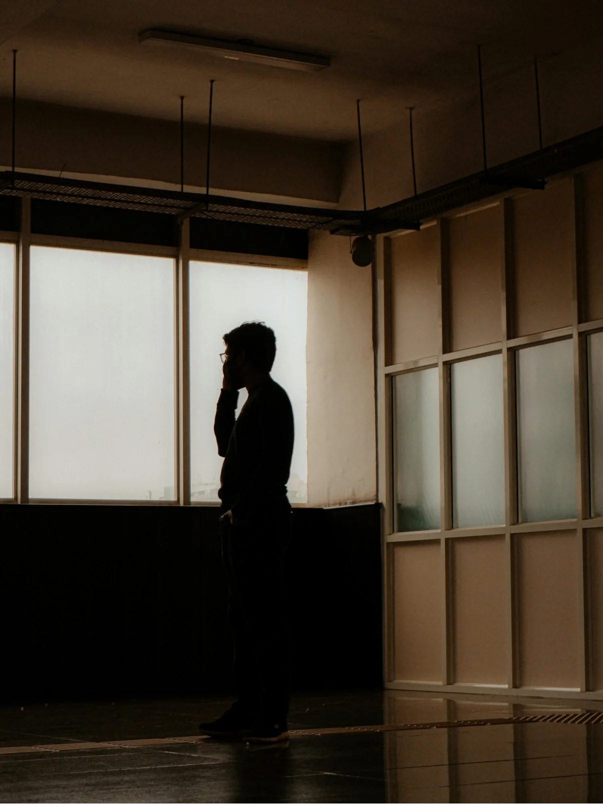 Silhouette of a man standing indoors near a large window, talking on the phone, with a modern office interior and frosted glass panels.