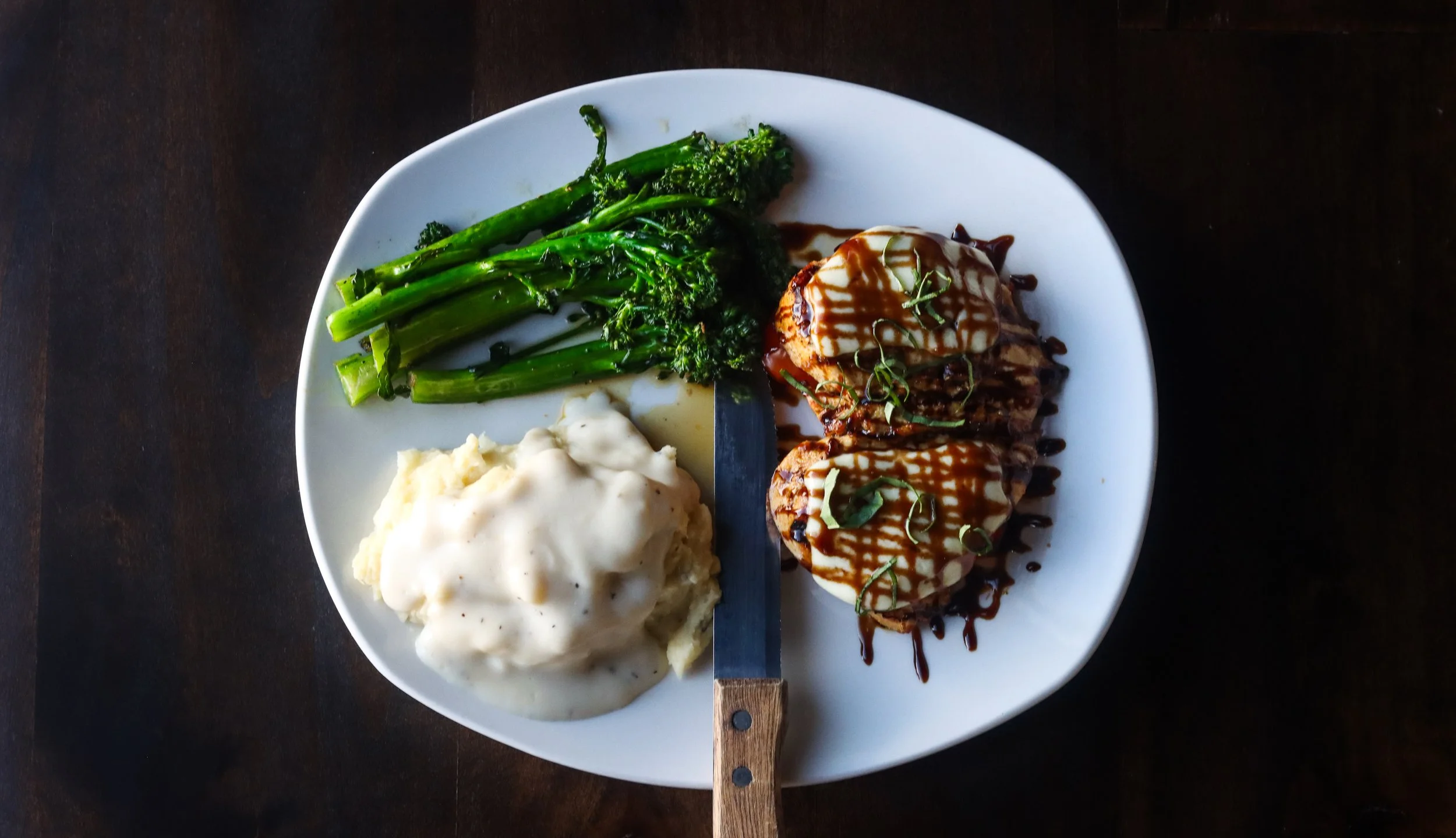 Plate with green vegetables, mashed potatoes with gravy, and grilled steaks topped with sauce and garnished with herbs.