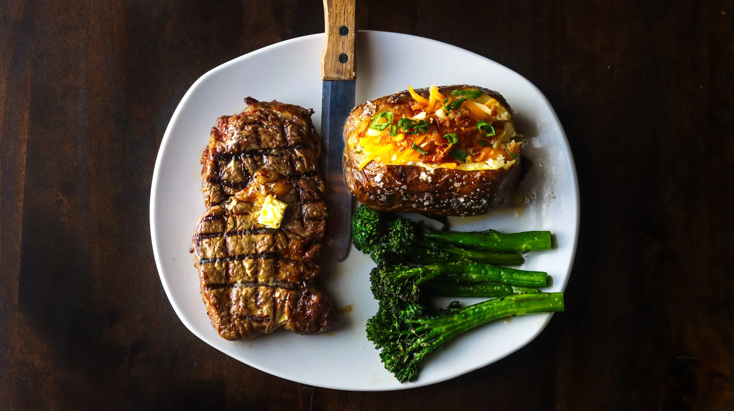 A white plate with grilled steak, baked potato topped with cheese and green onions, and steamed broccoli on a dark wooden surface.