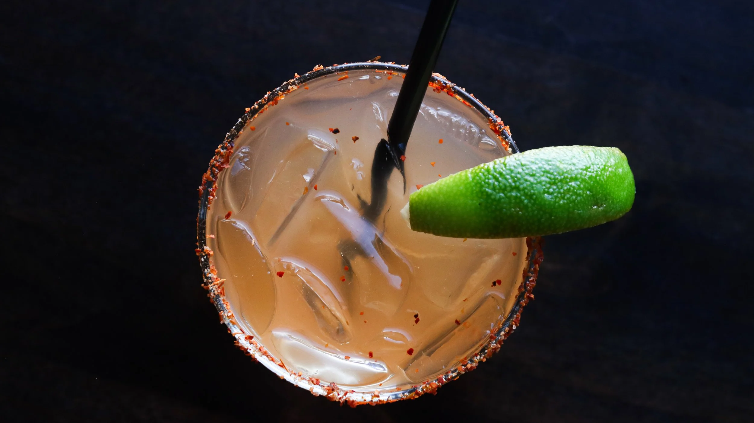 Top-down view of a cocktail glass with a salted rim, filled with ice and a brownish drink, garnished with a lime wedge, on a dark surface.