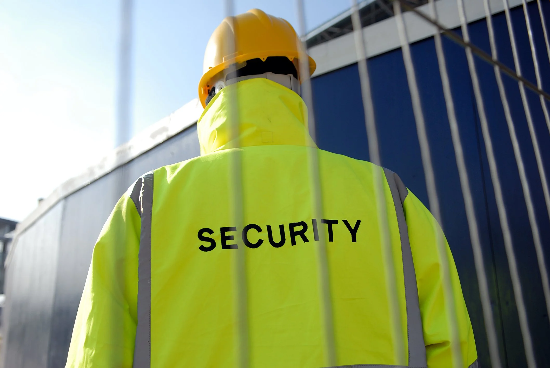 Security guard in a yellow jacket and helmet standing outside behind a metal fence