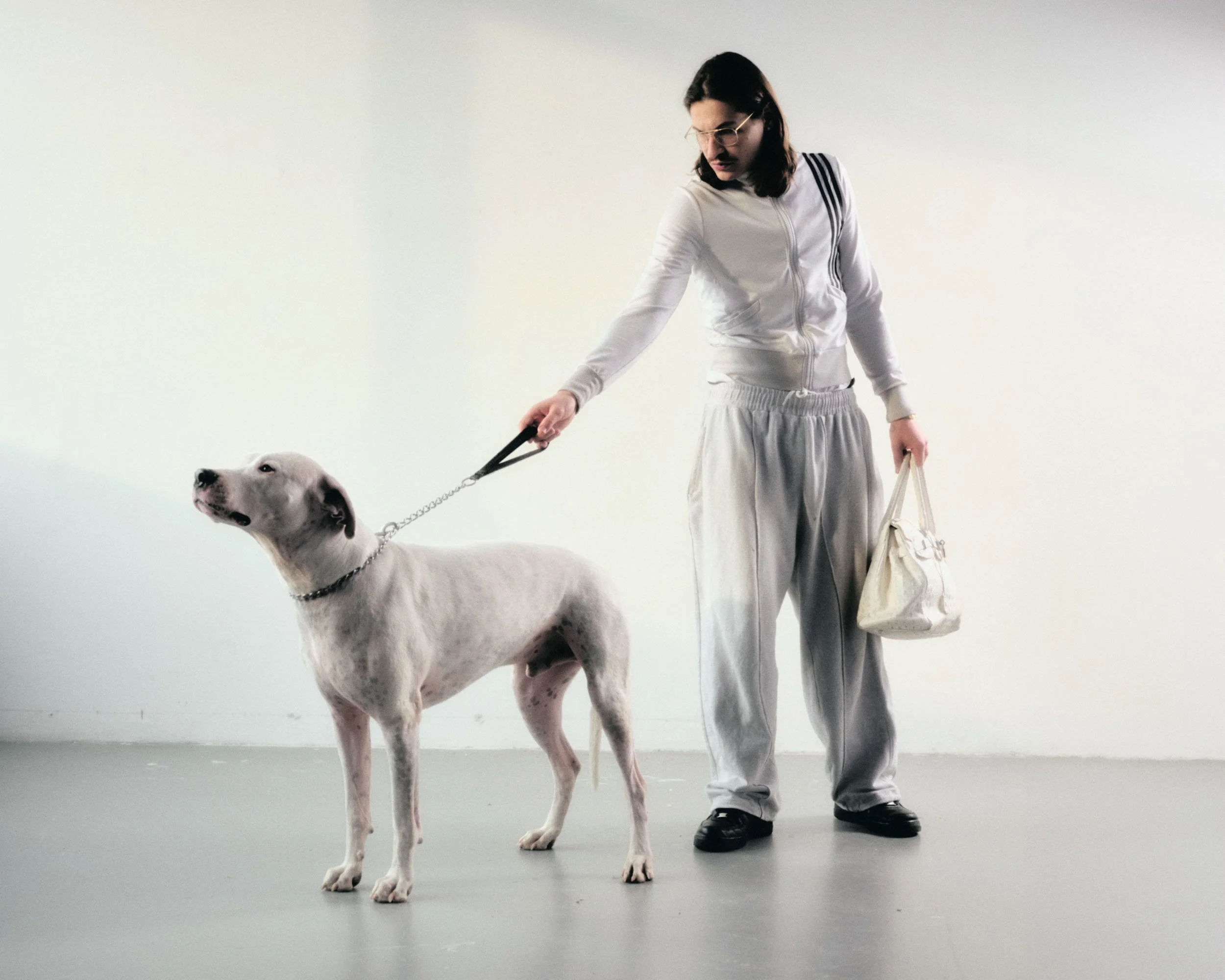 Eine Frau mit dunklen Haaren in sportlicher Kleidung hält eine Leine an einem weißen Hund mit schwarzen Flecken. Die Frau trägt eine weiße Jacke, weite graue Hosen und schwarze Schuhe, und hält eine weiße Tasche in der Hand.