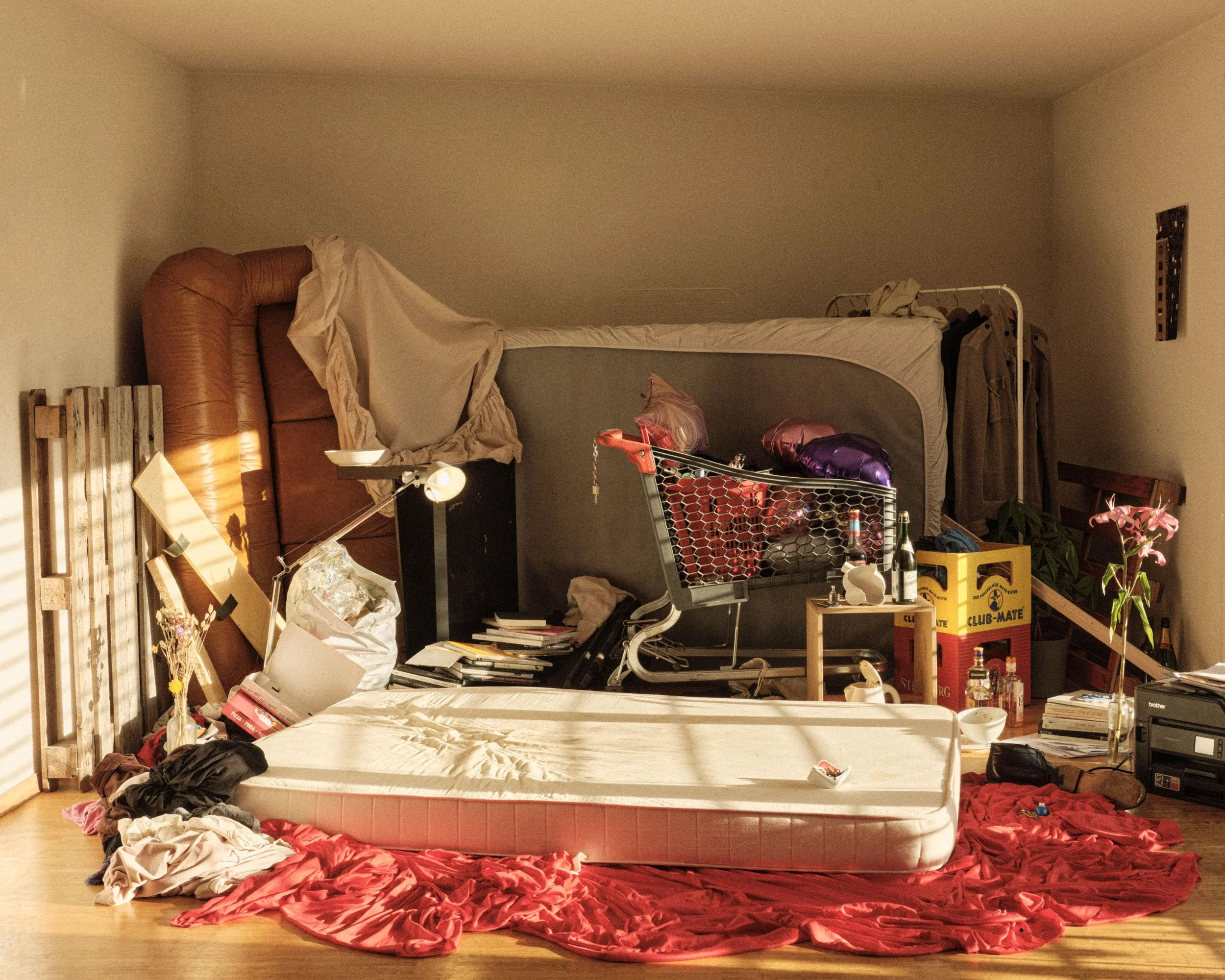 Unordentliches Zimmer mit umgestürztem Matratze auf einem roten Bettlaken, verschiedene Gegenstände, Bücher, leere Flaschen, eine Einkaufskarre mit Luftballons, Kleidung, Pflanzen und Möbel.