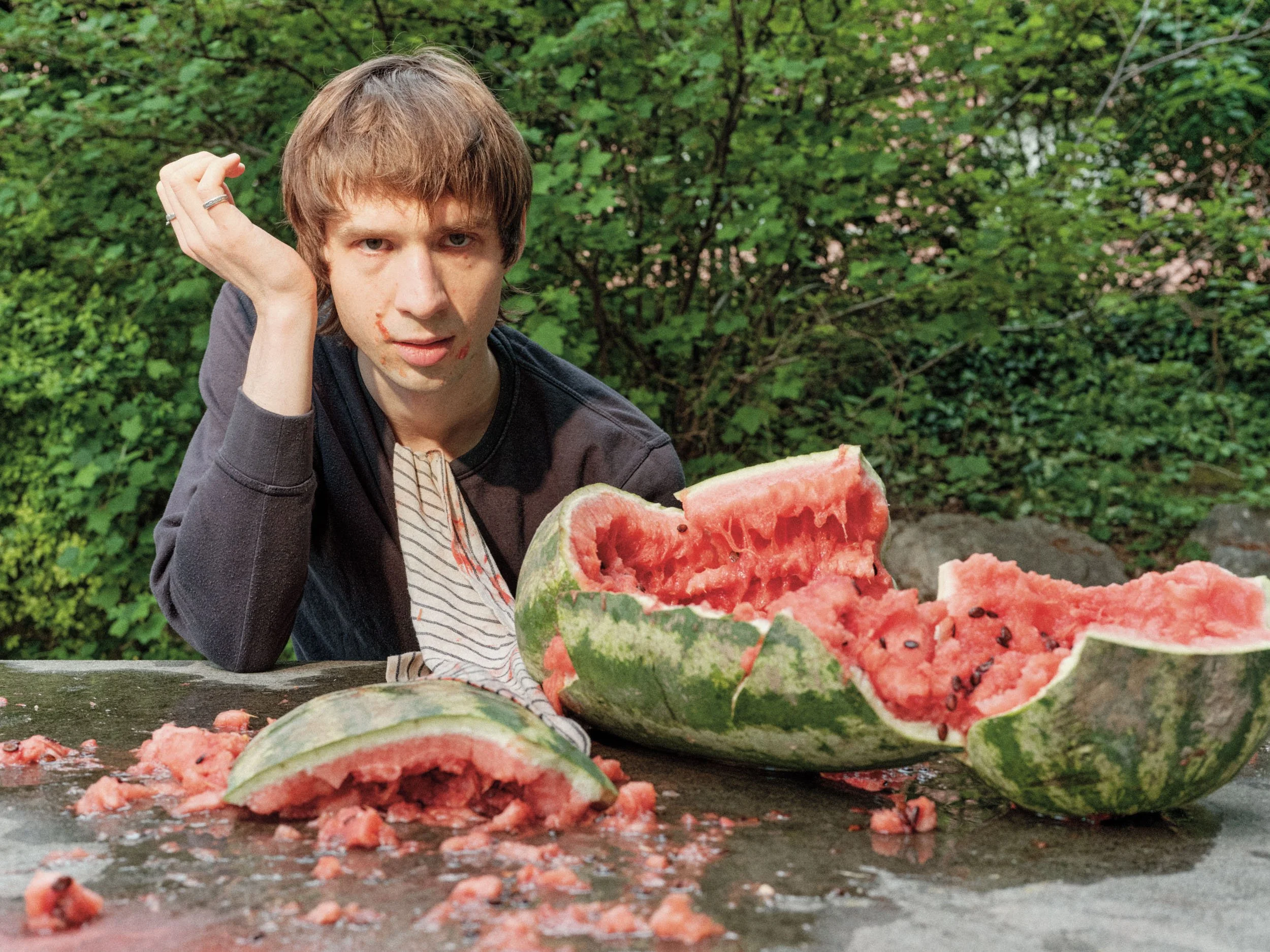 Ein junger Mann mit braunen Haaren und leichter Verletzung im Gesicht sitzt vor einer aufgeschnittenen Wassermelone, die viele schwarze Samen enthält, im Freien mit grünen Büschen im Hintergrund.