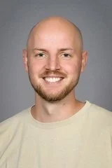 Portrett av en mann med lyst hår, skjegg og smil, mot en nøytral grå bakgrunn.