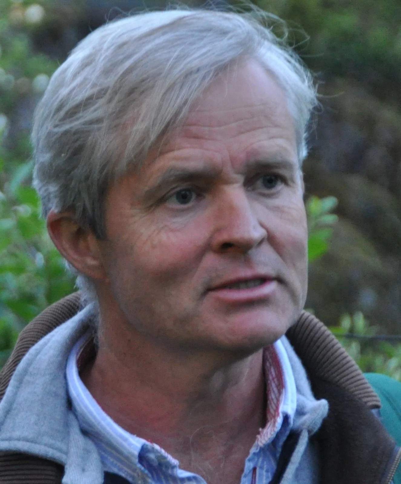 Et portrett av en mann med blond hår, iført skjorte og jakke, ute i naturen med grønne blader i bakgrunnen.
