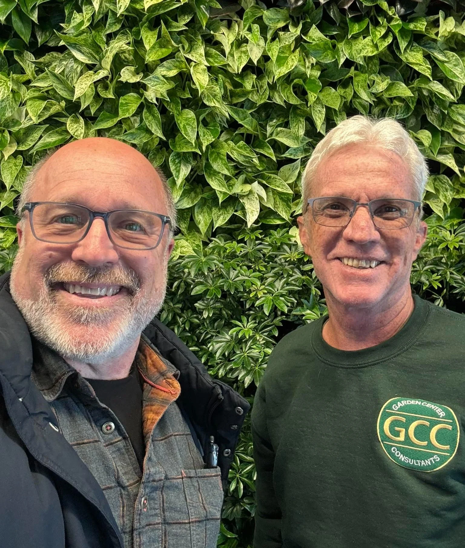 Two smiling men in glasses standing in front of a green leafy wall, one wearing a dark jacket and plaid shirt, the other in a dark green shirt with a company logo.