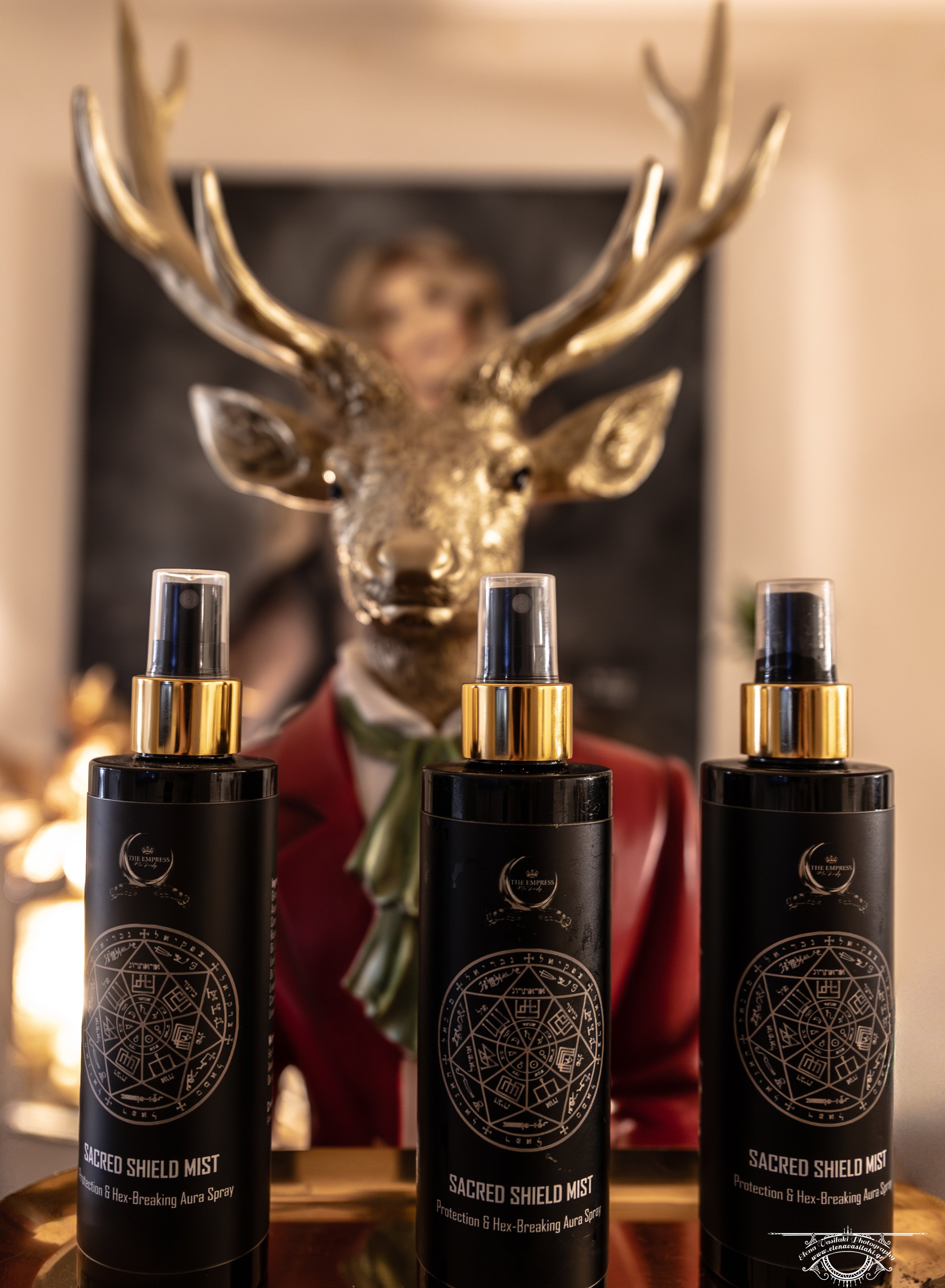 Three black sprays labeled 'Sacred Shield Mist' with gold caps in the foreground, and a gold deer head sculpture and a blurred portrait of a woman in a red blazer in the background.
