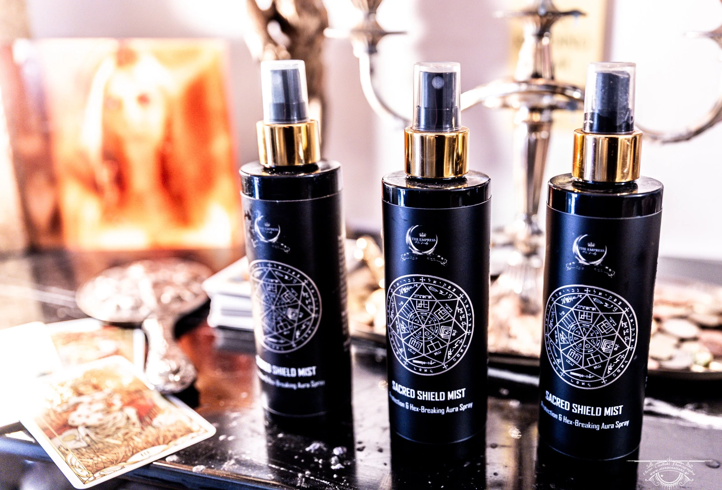 Three black bottles labeled Sacred Shield Mist with gold spray tops on a black surface, with tarot cards and other objects in background.