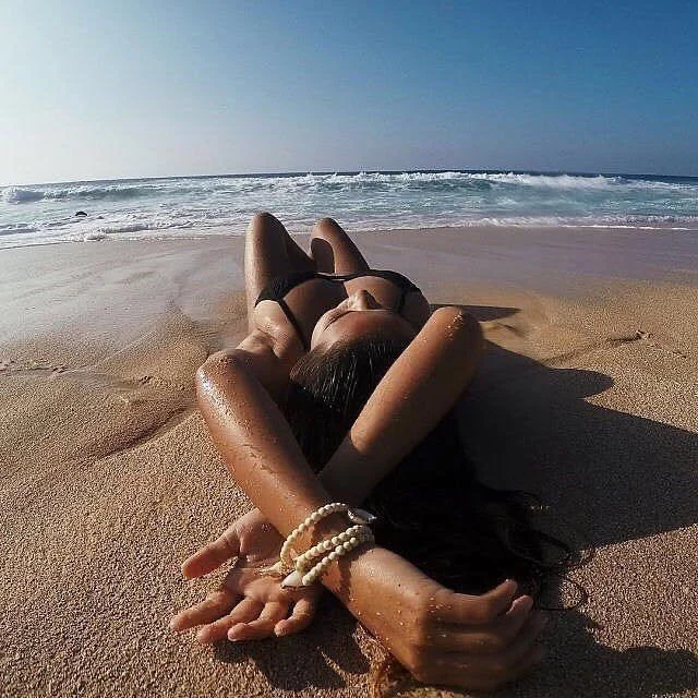 Person lying on sandy beach in a black swimsuit, with arms behind head and bracelets on wrist, facing the ocean under a clear sky.