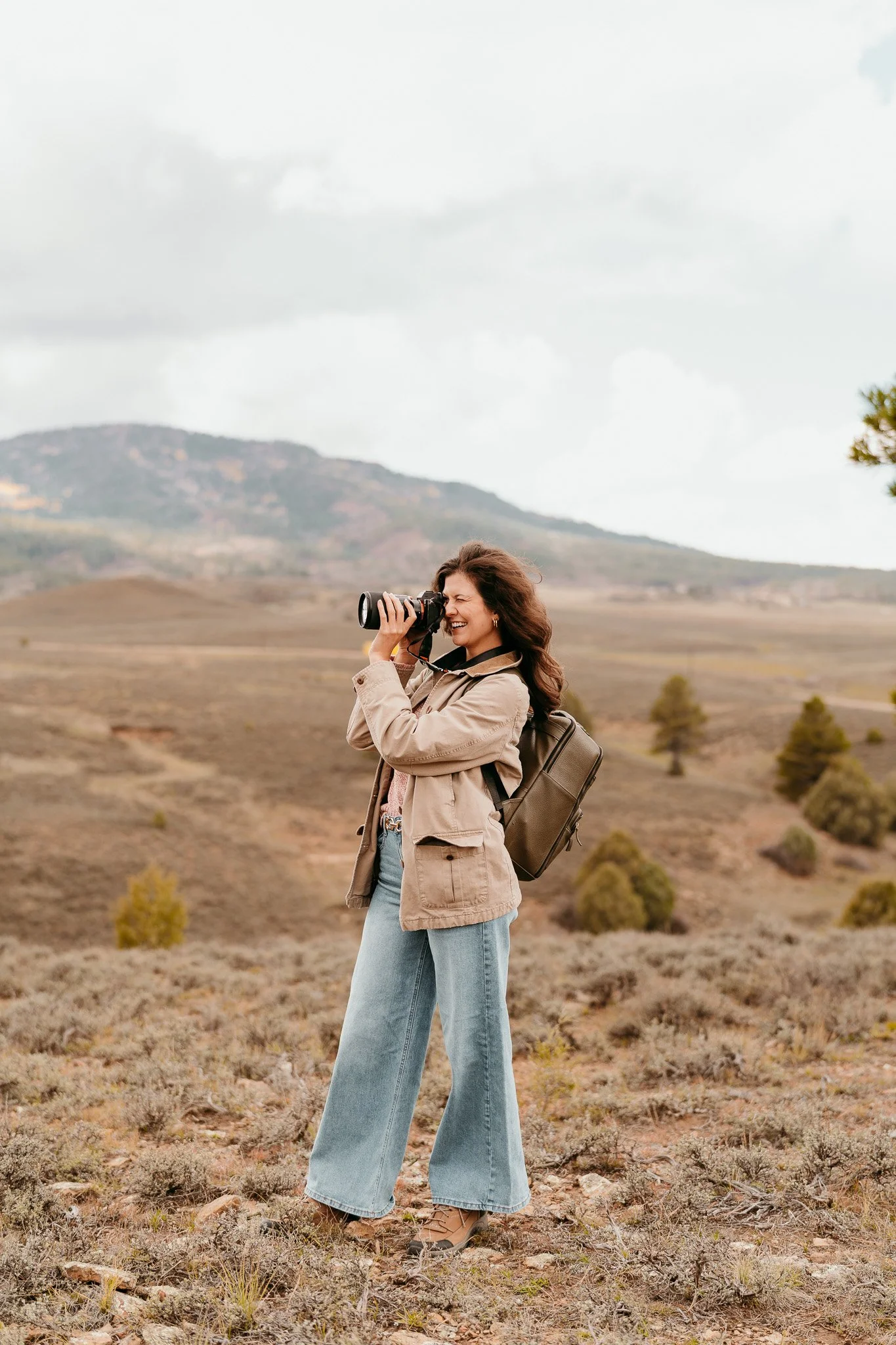 A woman in beige jacket and blue jeans standing in a landscape with rolling hills, holding a camera to her eye and smiling.