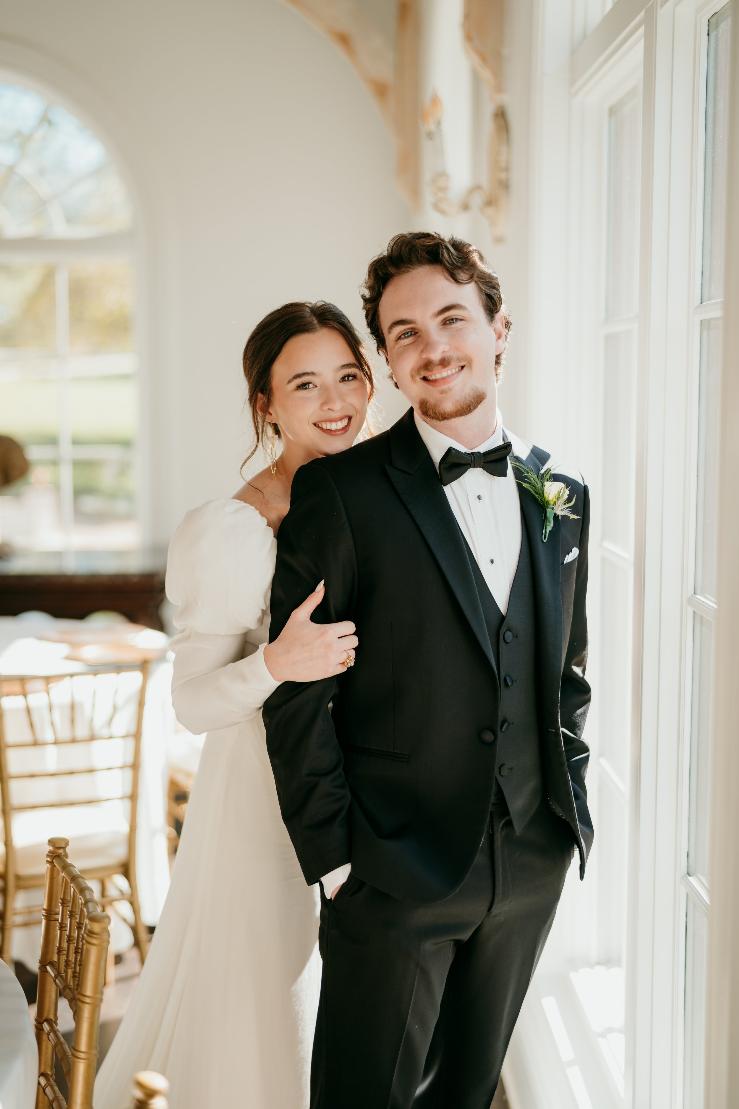 A bride and groom standing near a sunlit window, smiling for a wedding photo. The bride is wearing a white wedding dress, and the groom is dressed in a black tuxedo with a bow tie, boutonniere, and pocket square.