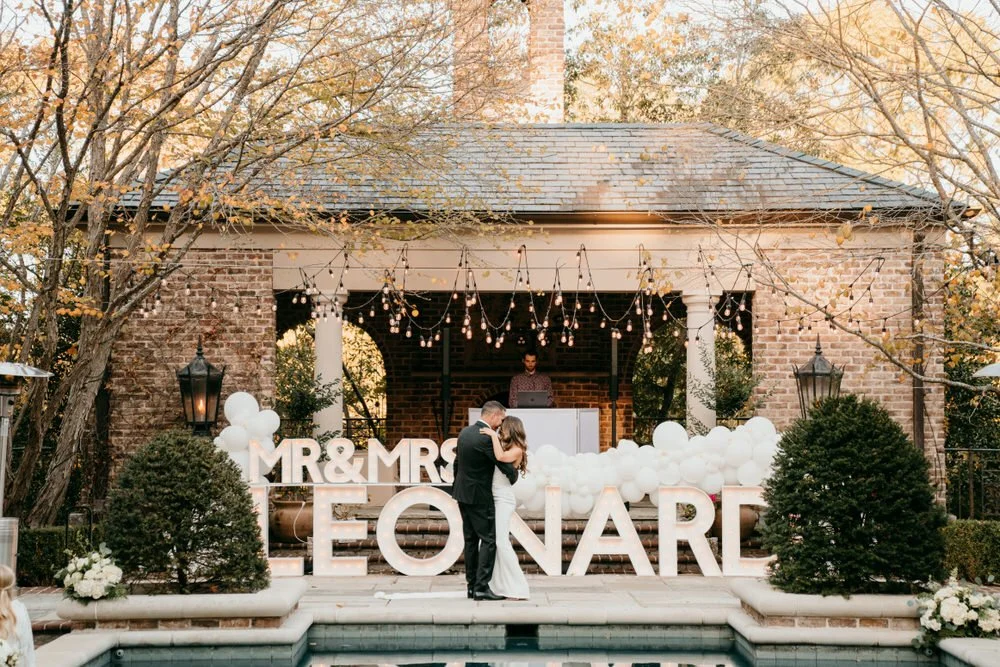 A newlywed couple dancing in front of a decorated outdoor wedding stage with large letters spelling 'Mr & Mrs Leonard', surrounded by balloons and string lights, with a DJ in the background.