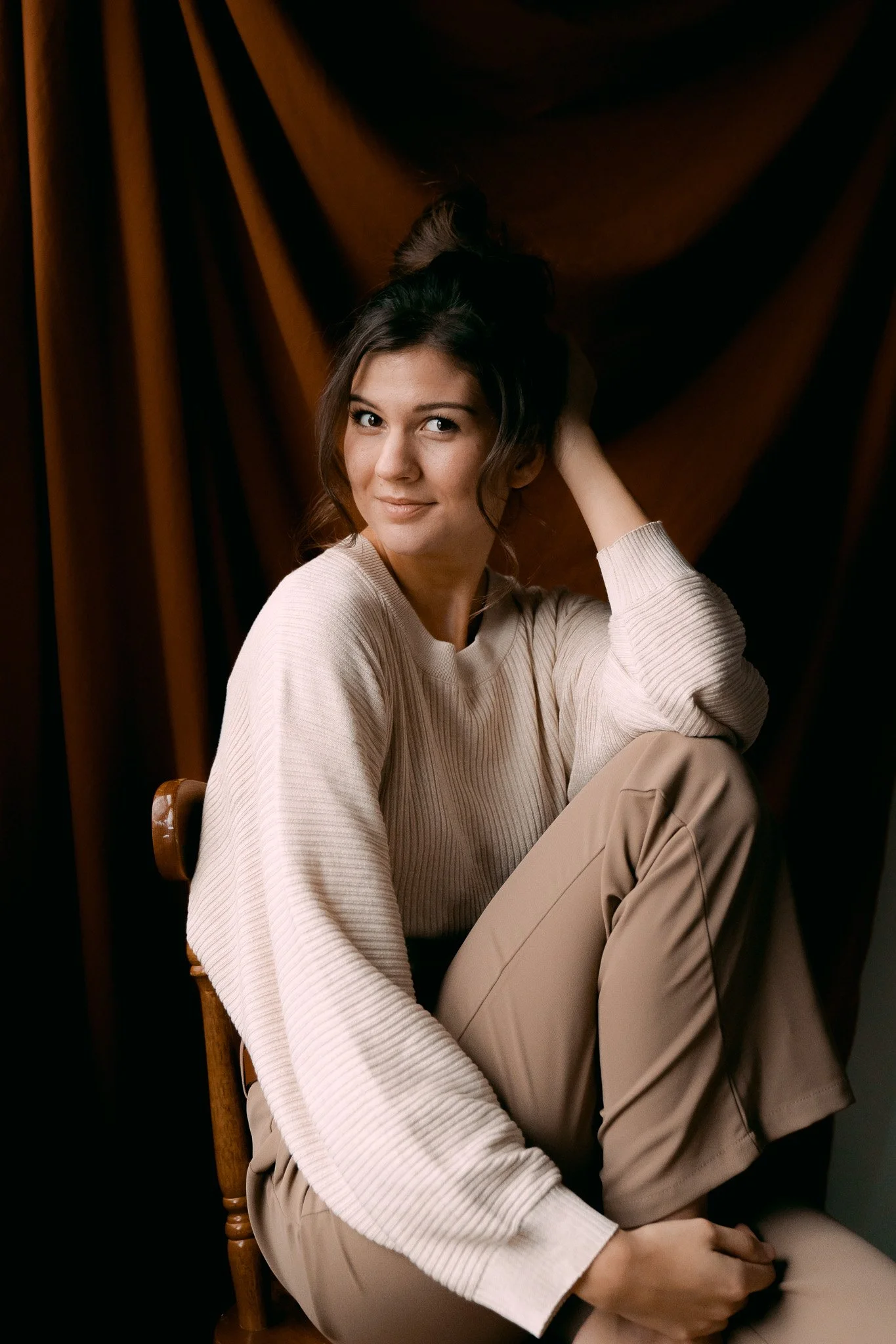 A young woman sitting on a chair, wearing a beige sweater and pants, with dark hair tied up, against a dark brown curtain background.