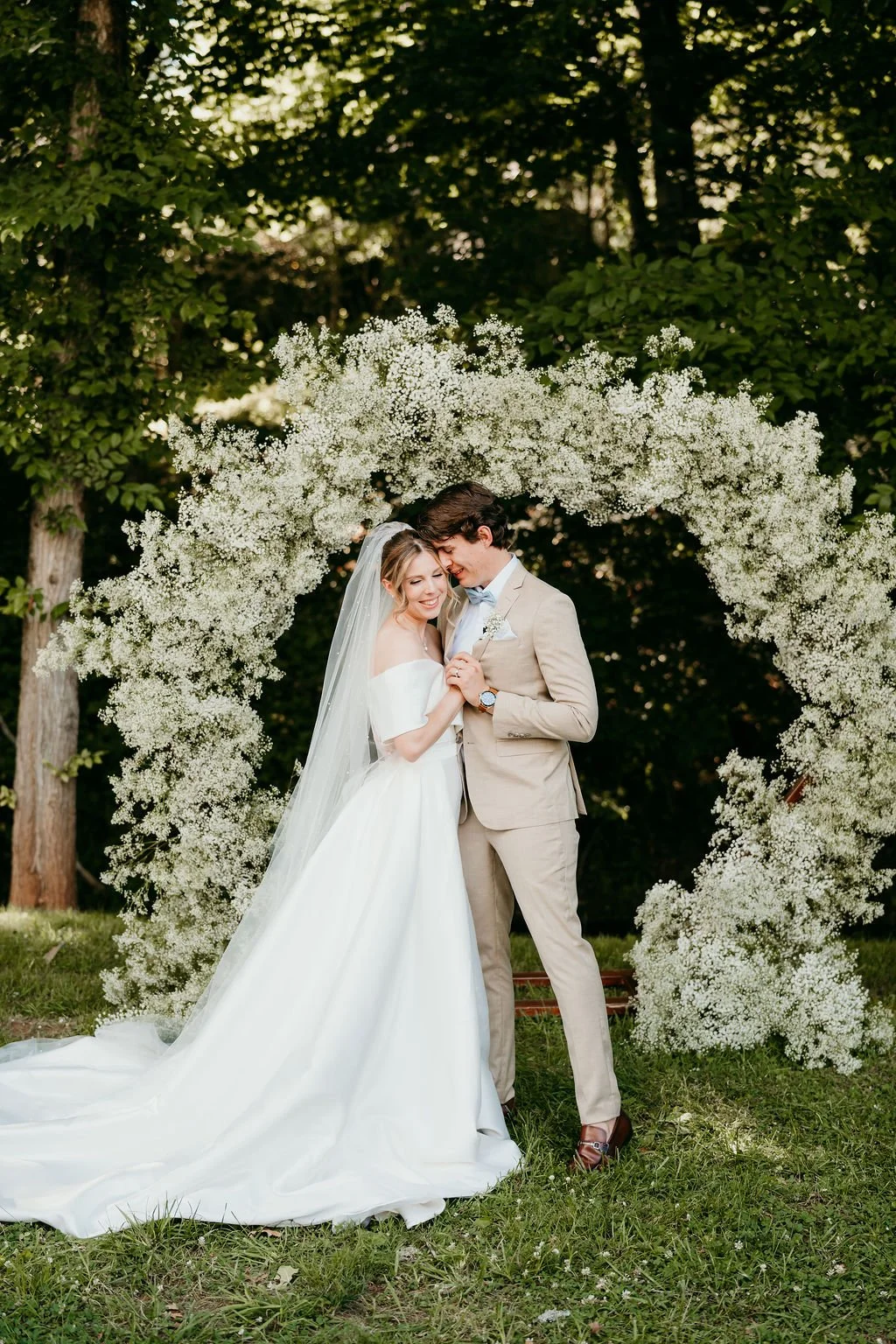 A bride and groom standing under a white floral arch outdoors during their wedding, smiling and embracing each other.