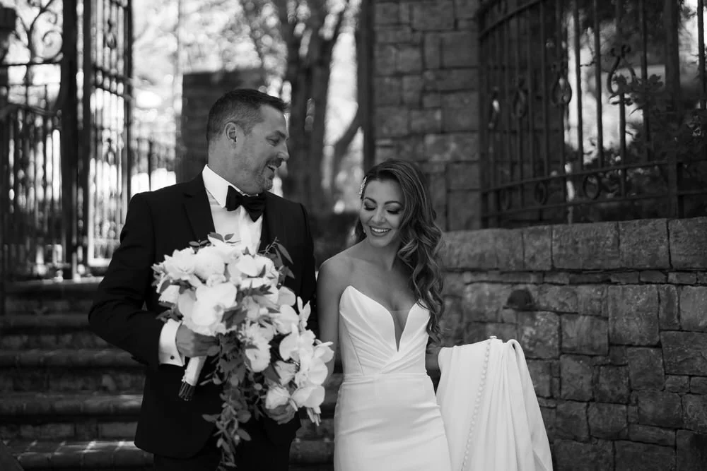 Black-and-white wedding photo of a groom in a tuxedo holding a bouquet and a smiling bride in a strapless gown carrying her bridal train.
