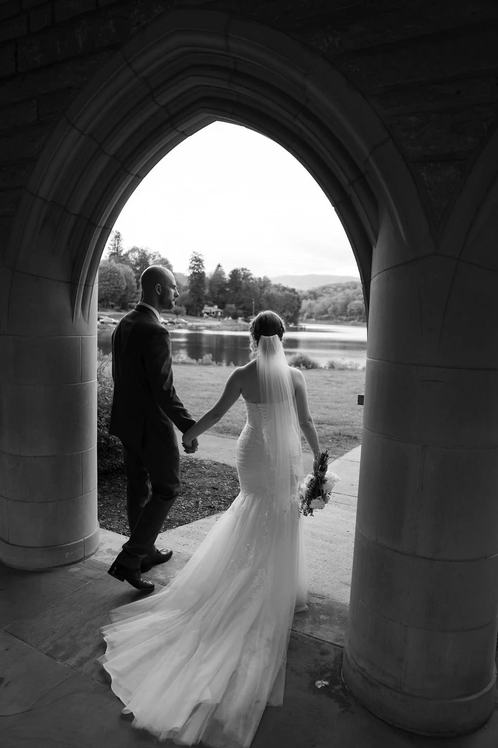 A bride and groom holding hands and walking down the aisle at an outdoor wedding, surrounded by seated guests and trees.