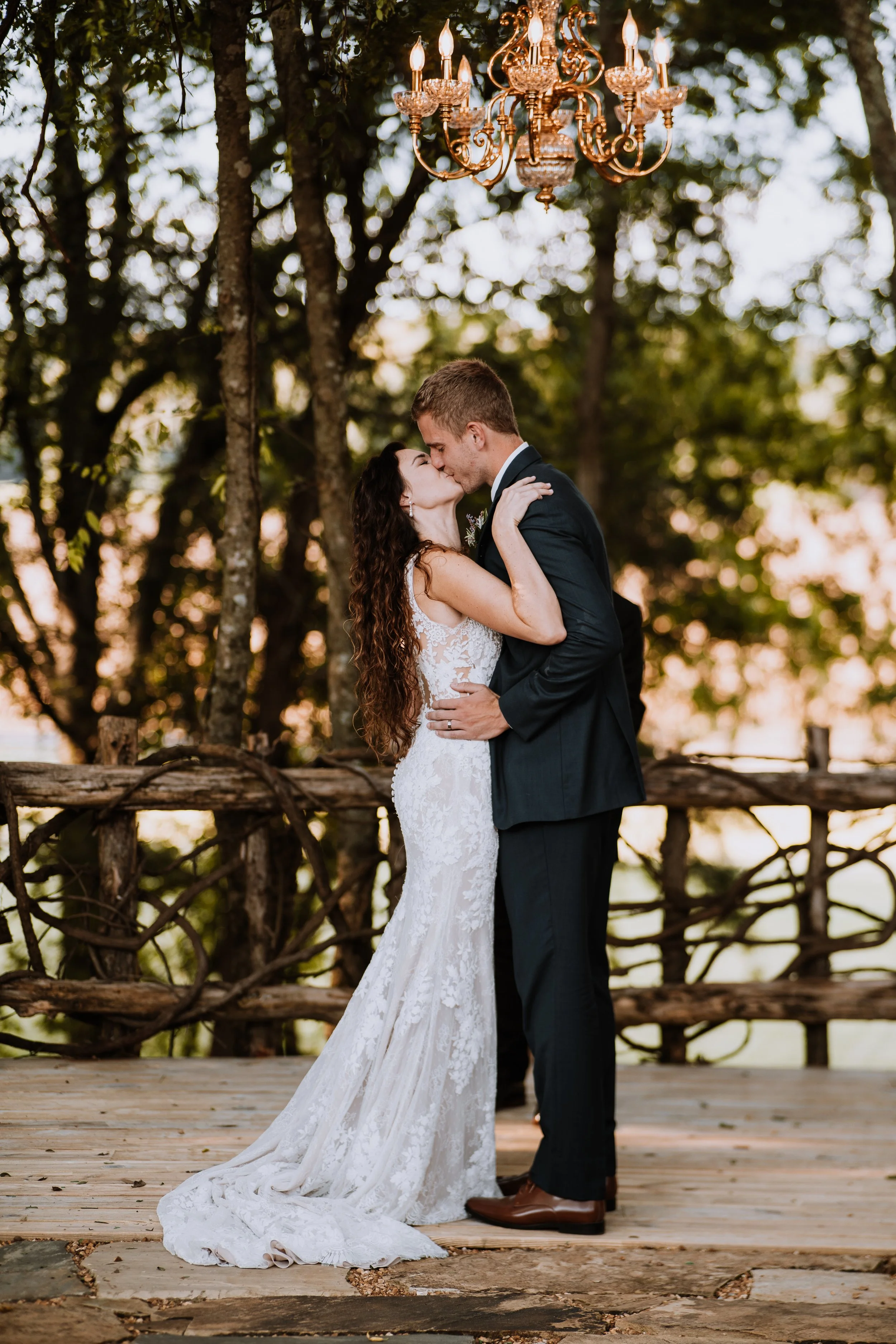 A bride and groom share a kiss outdoors during sunset, with a chandelier hanging above them and a rustic wooden fence in the background.