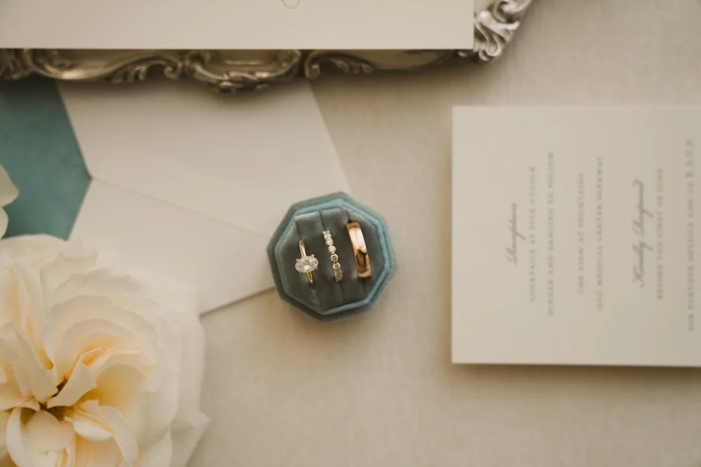 A set of three rings, including a solitaire engagement ring with a diamond, a diamond wedding band, and a plain gold ring, displayed in a small blue velvet ring box on a table with wedding invitations, flowers, and a decorative mirror border.
