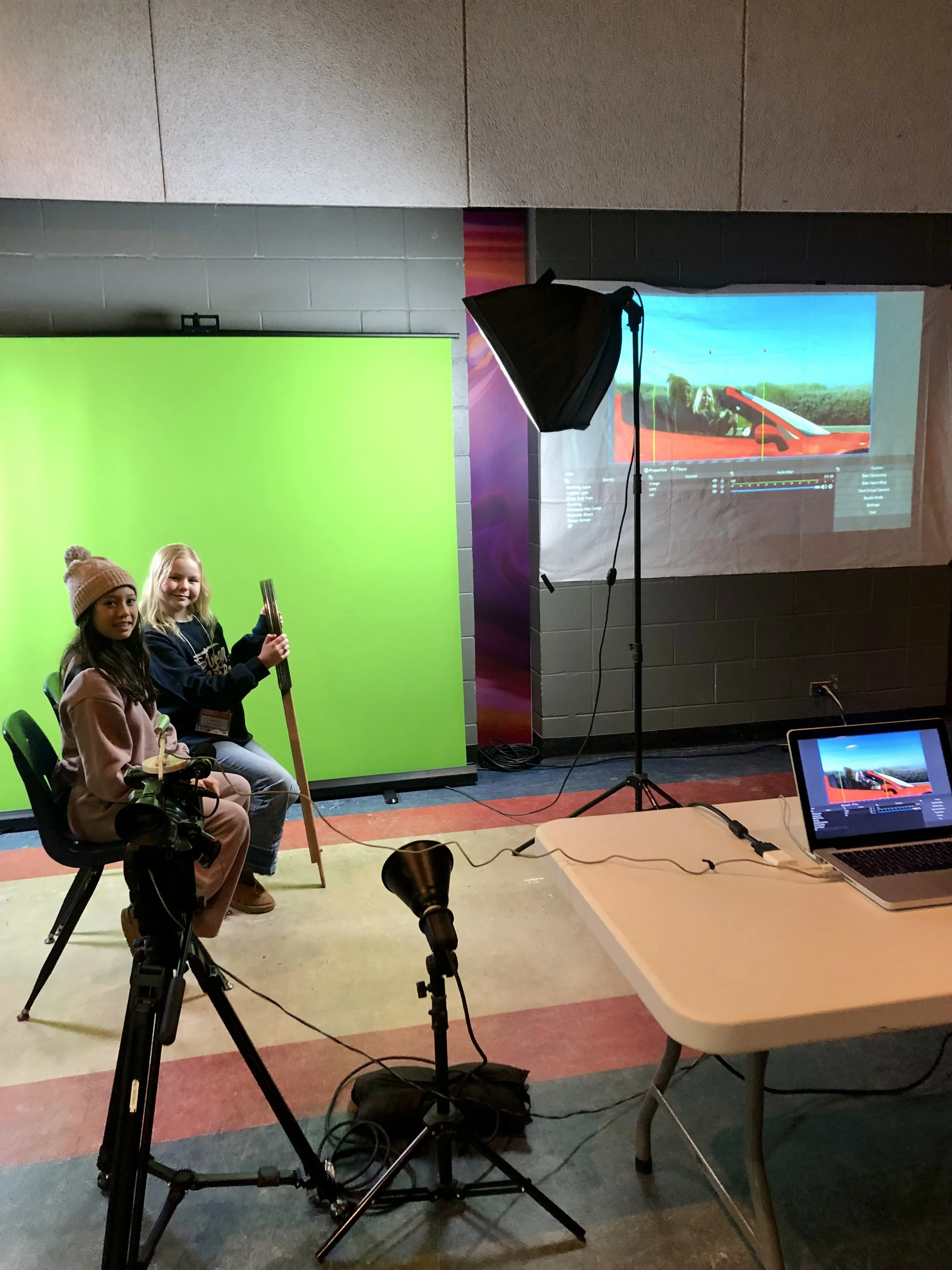 Two girls in front of a green screen with a projection of them driving to the right
