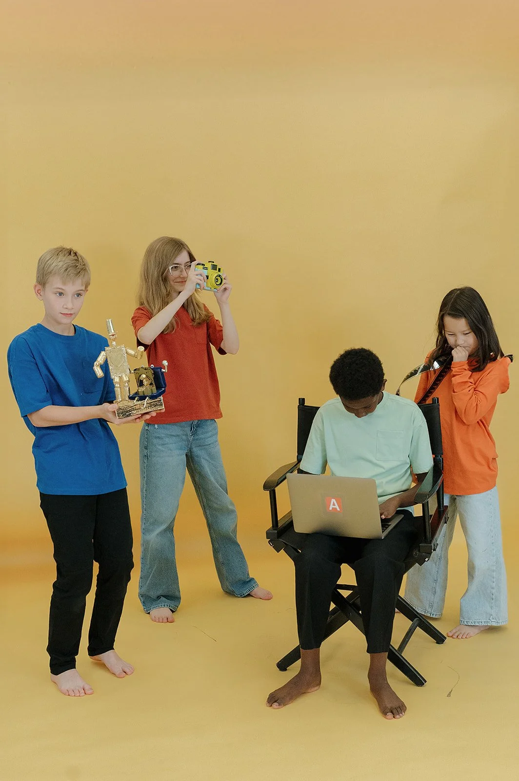 Four kids, a boy holding a trophy, a girl with a camera, a boy sitting on a director's chair on a laptop and a girl holding a film strip