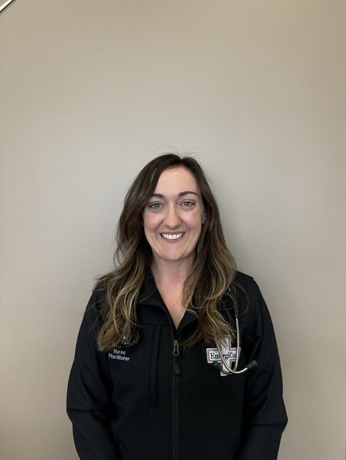 A woman with wavy brown hair smiling at the camera, wearing a black jacket with a name tag and a stethoscope clipped to her jacket, standing against a plain beige wall.