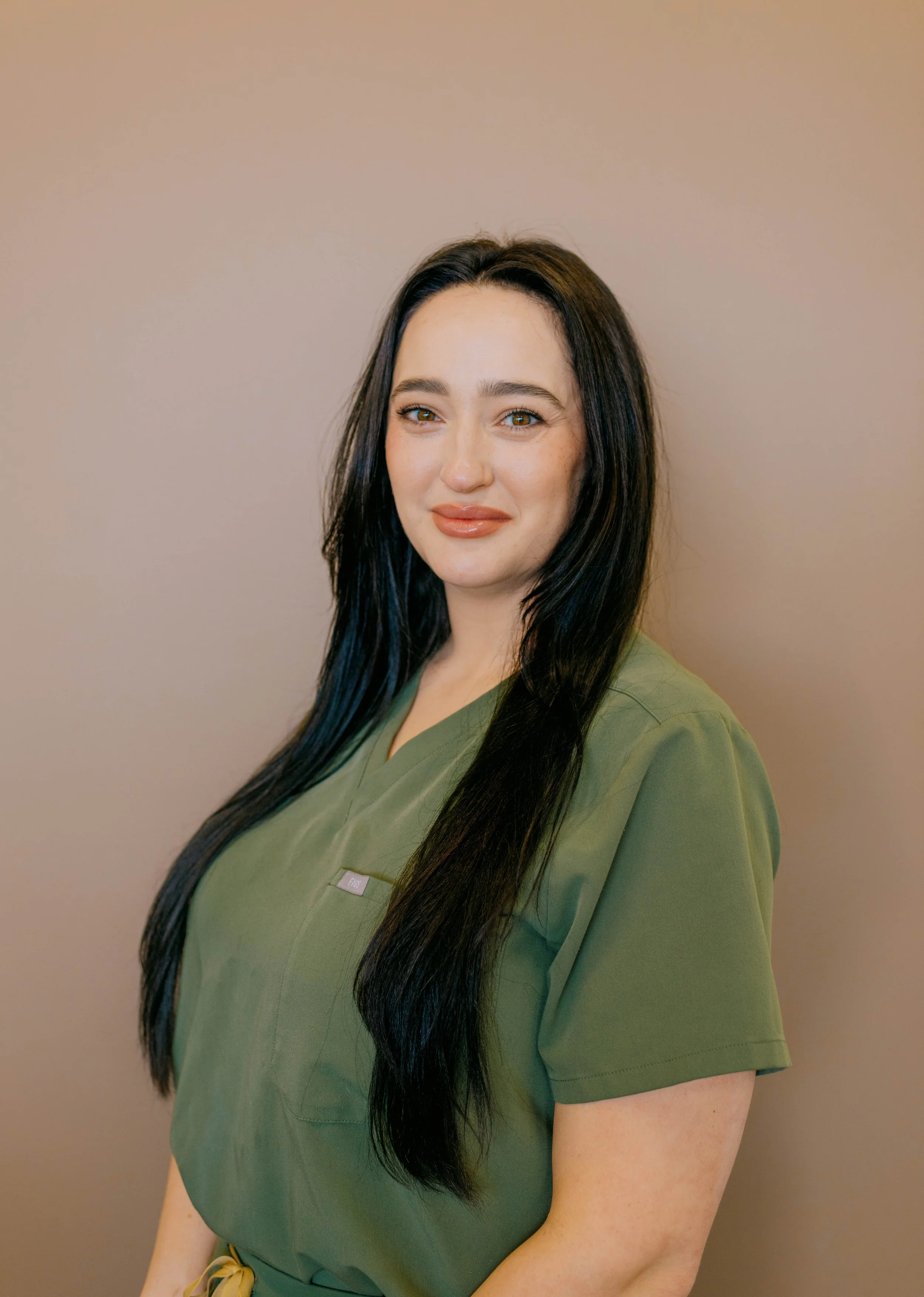 A woman with long dark hair, light skin, and wearing an olive green scrub top, standing against a plain beige wall.