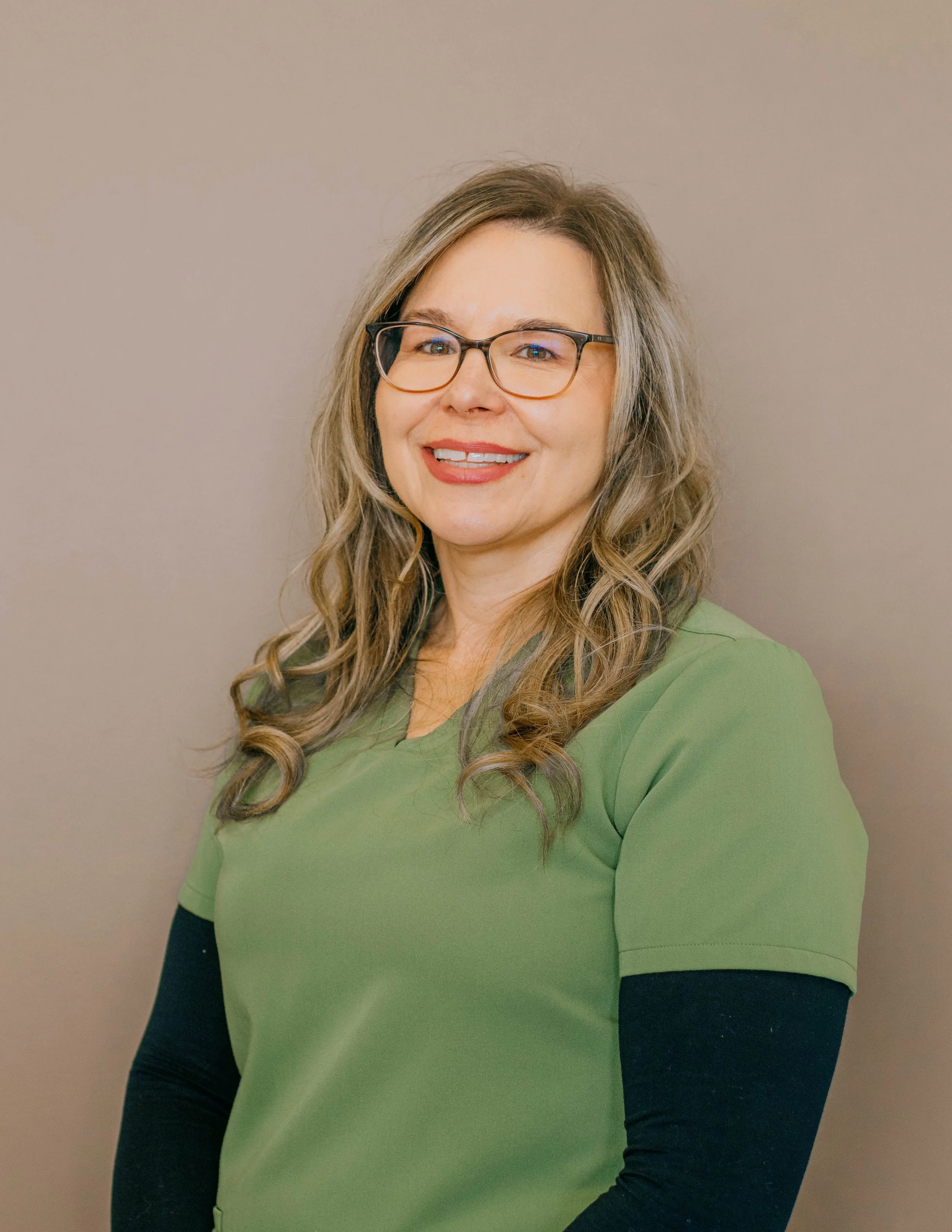 A woman with long wavy blonde hair, glasses, and a friendly smile, wearing a green top with black sleeves, standing against a plain beige wall.