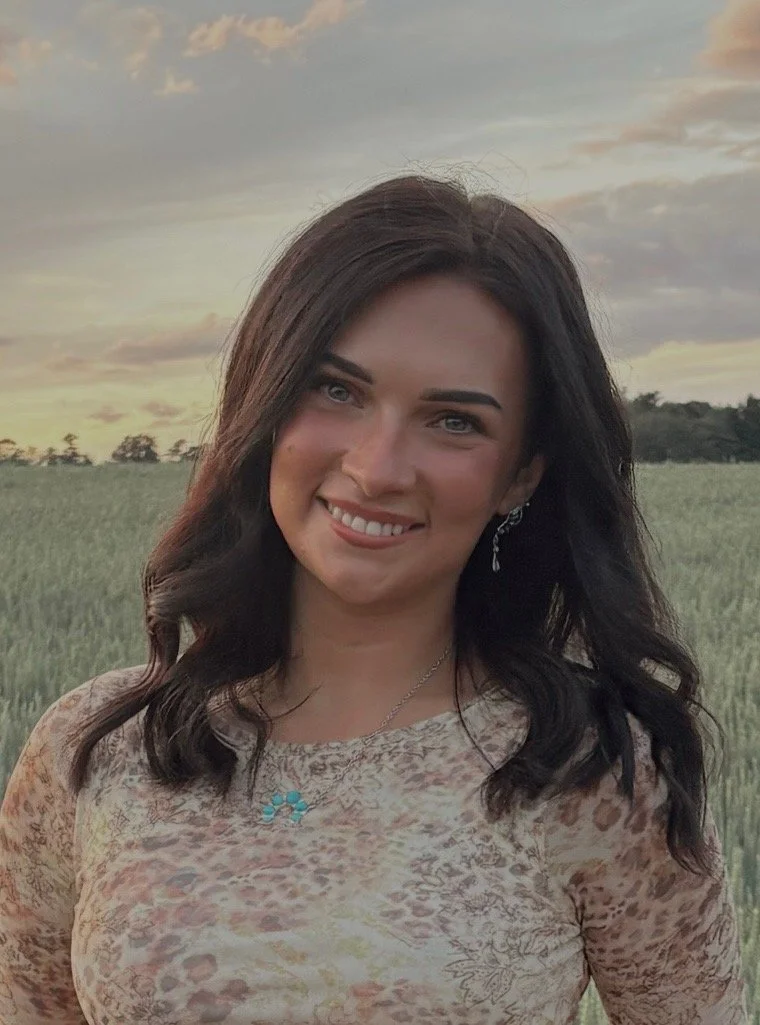 A woman with dark hair smiling outdoors during sunset, standing in a field of tall grass. She is wearing a patterned top, earrings, and a necklace.