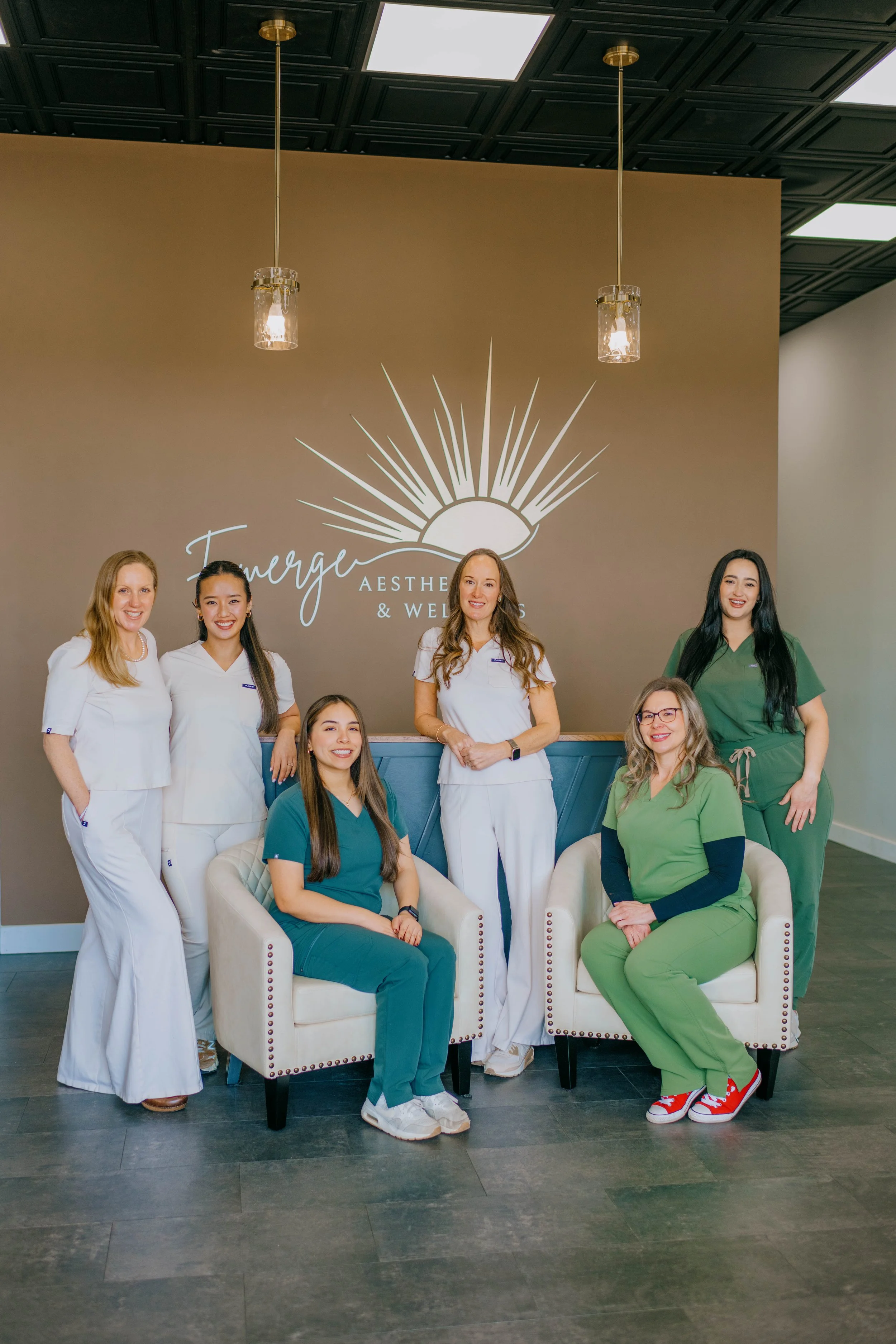 Group of seven women, some in medical scrubs and some in white uniforms, posing inside a modern aesthetic and wellness clinic. They are standing and sitting in front of a brown wall with a logo that reads 'Emurge Aesthetic & Wellness' under a stylized sun graphic. The setting features a black coffered ceiling with pendant lighting.