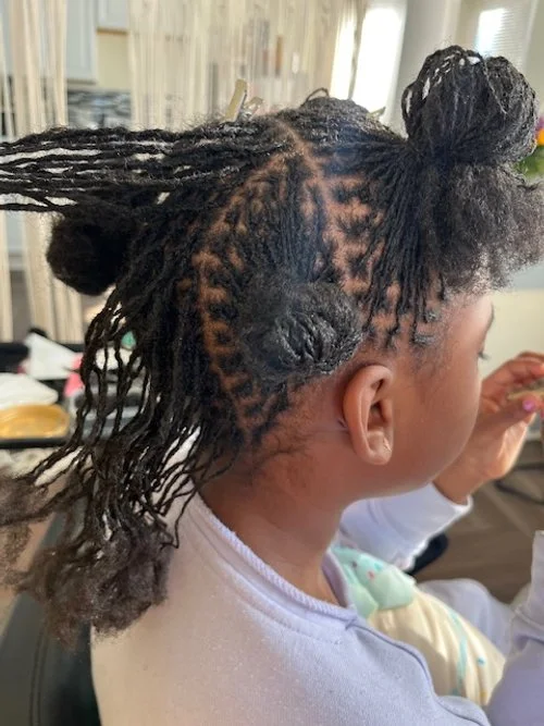 Child with partially styled curly hair, with the front section braided and two bun styles on top, sitting in a salon or home setting.