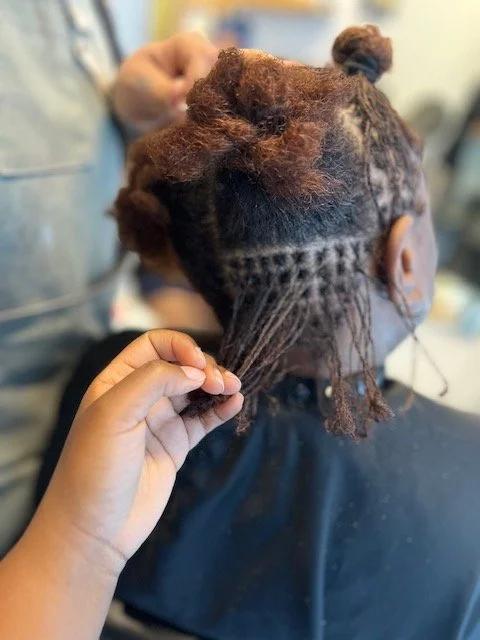 A person with partially styled hair, including a braided section and some loose strands, is having their hair twisted or styled by a stylist in a salon.