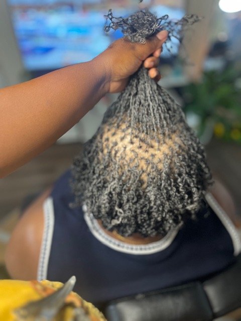 A person with partially styled hair, including a braided section and some loose strands, is having their hair twisted or styled by a stylist in a salon.